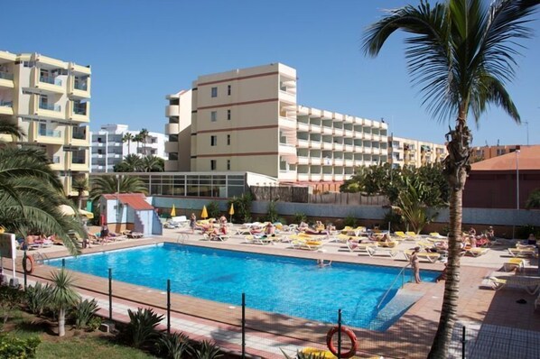 A swimming pool with sun loungers and a small poolside building, surrounded by palm trees and multi-story buildings.