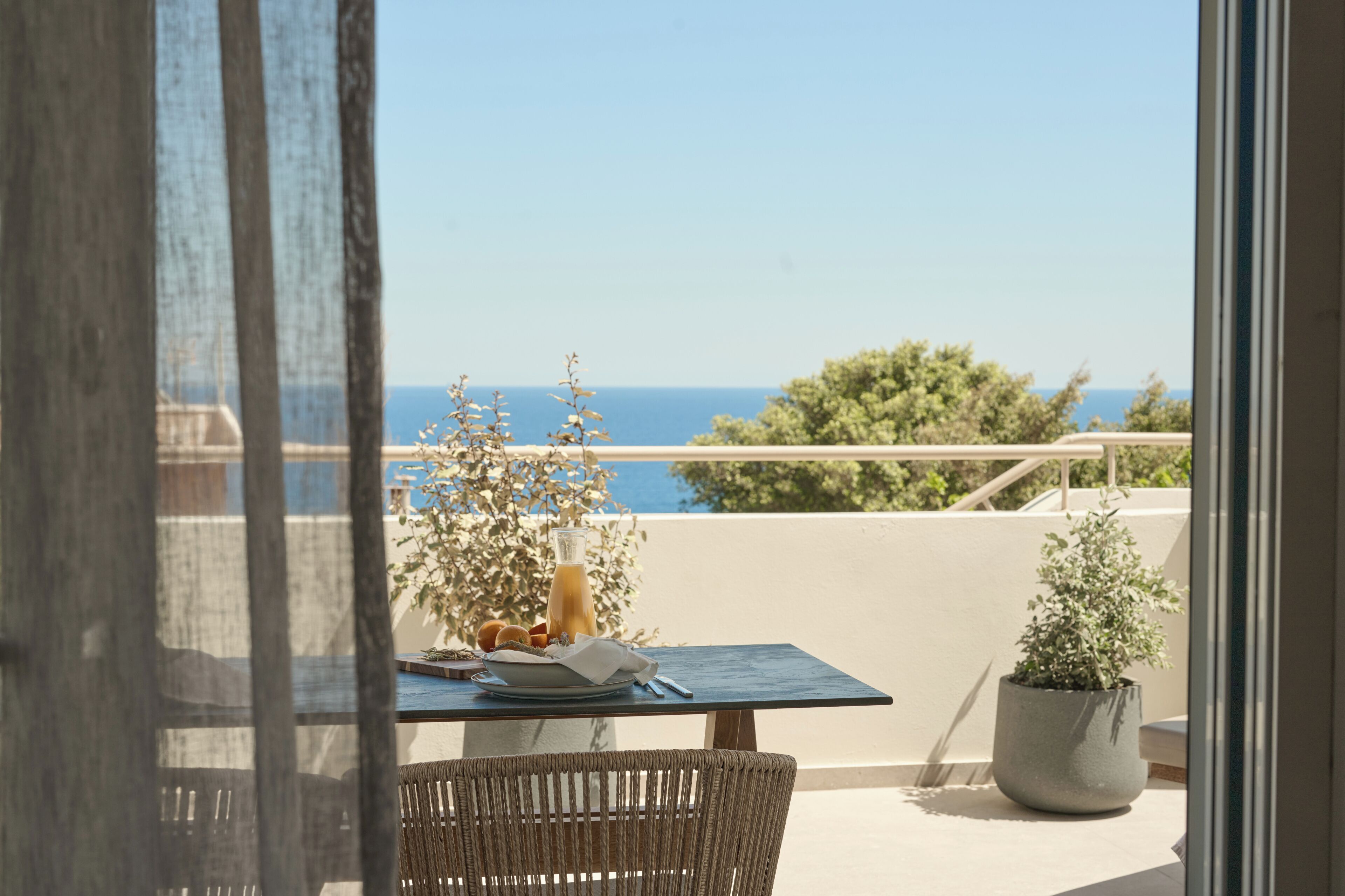 Un balcón con una mesa lista para el desayuno, una planta en maceta y vista al mar.