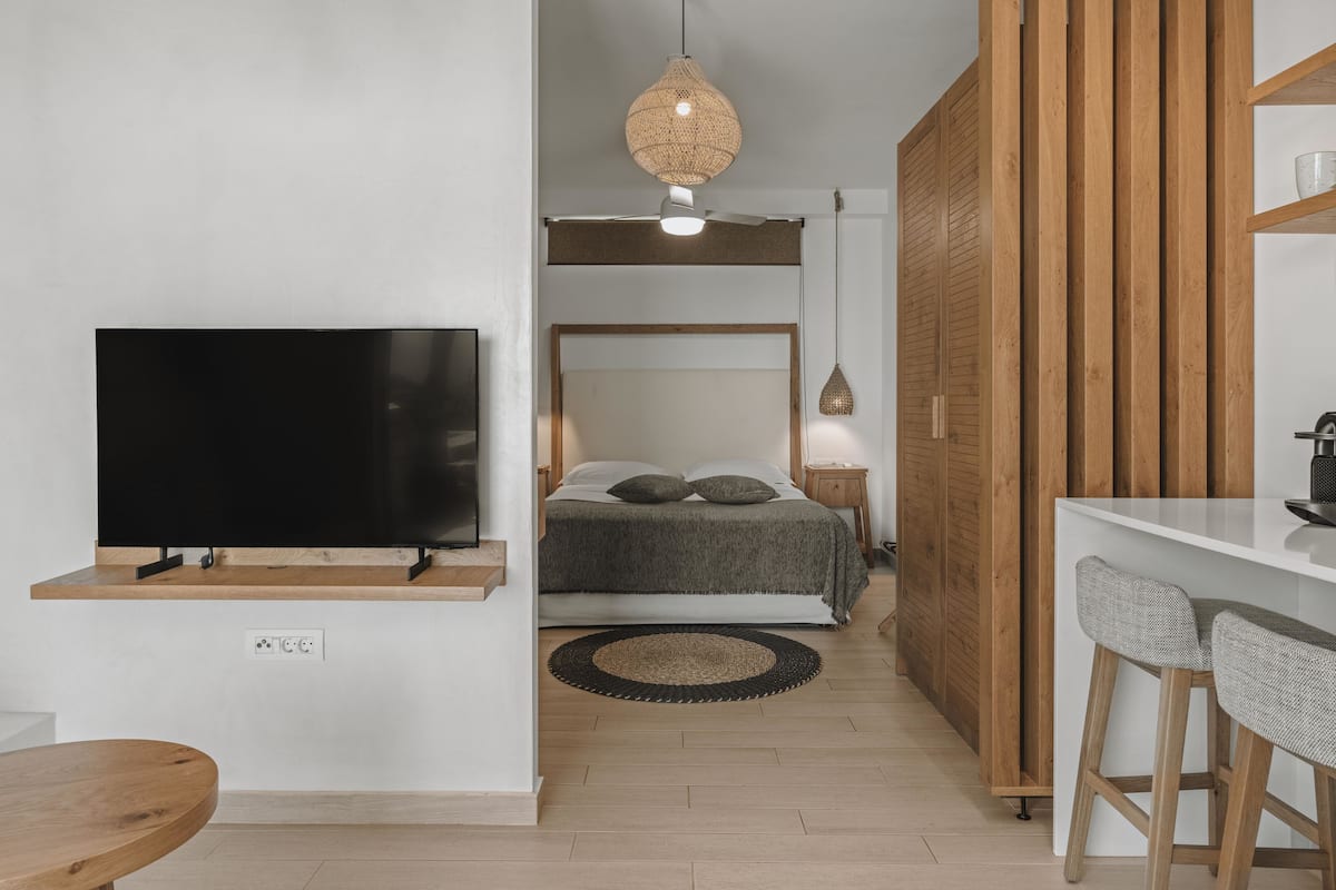 A compact living space with a TV mounted on the wall, a bed with a headboard, a small dining area with stools, and a wooden wardrobe.
