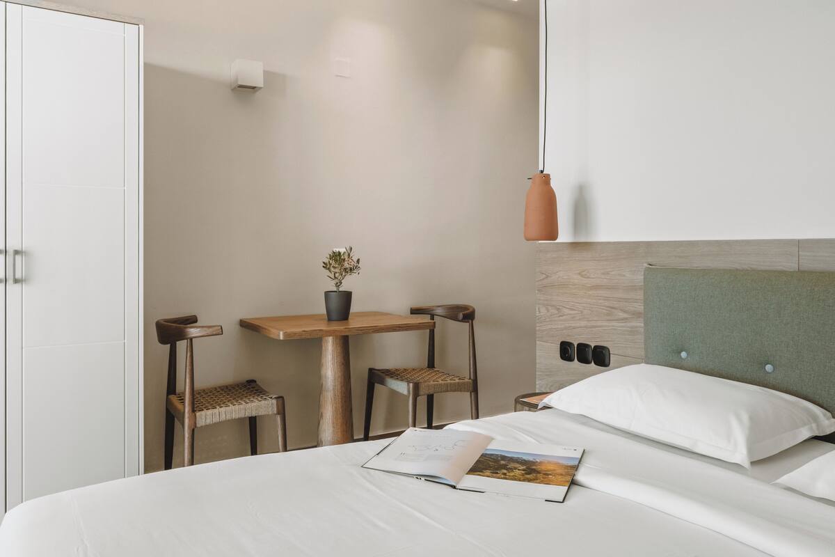 A hotel room with a bed, a desk with a chair, a small table with a plant, and a wall-mounted light fixture.