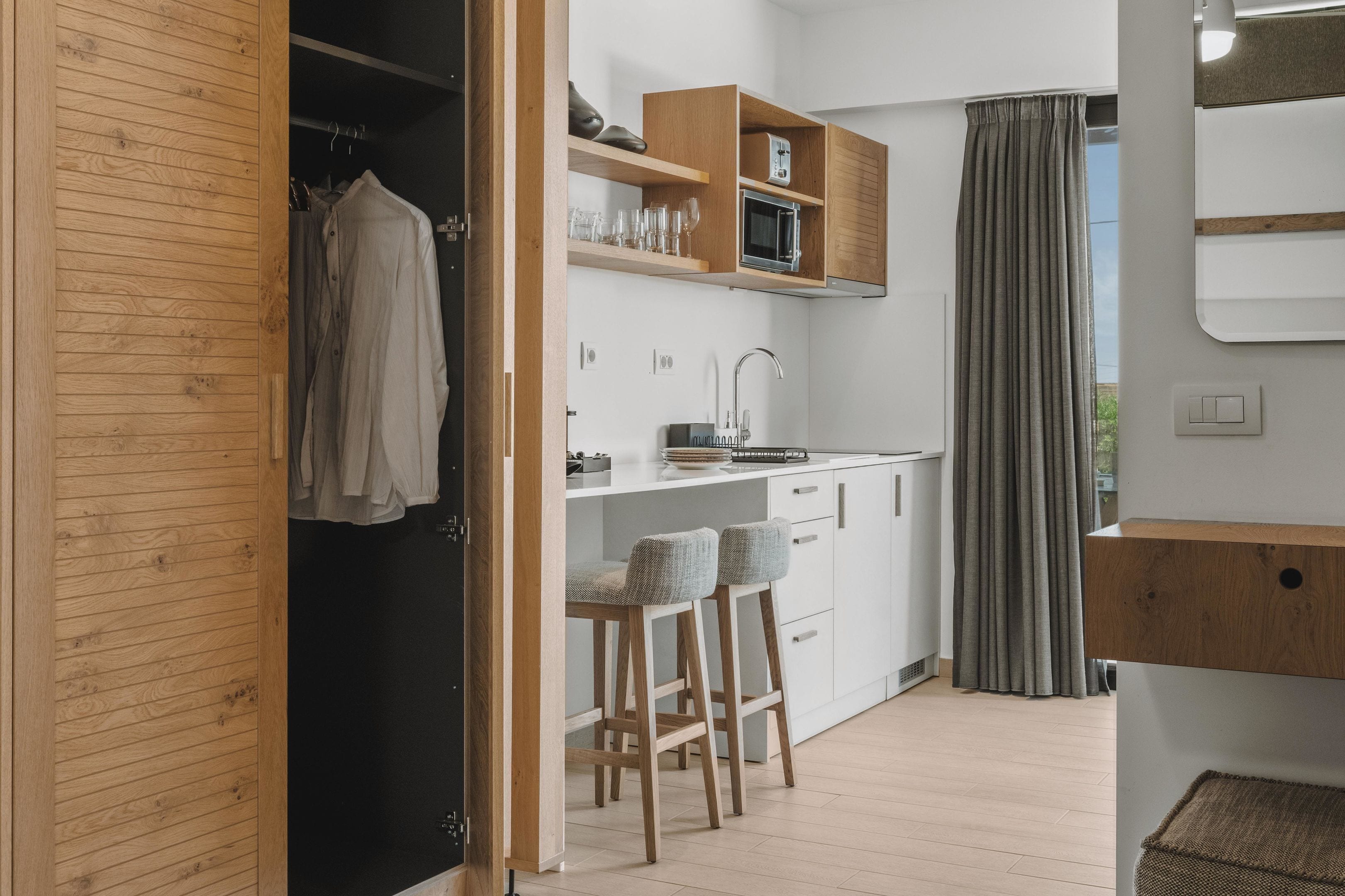 A compact kitchen with wooden cabinets, a microwave, and a sink. There are two bar stools and a window with curtains.