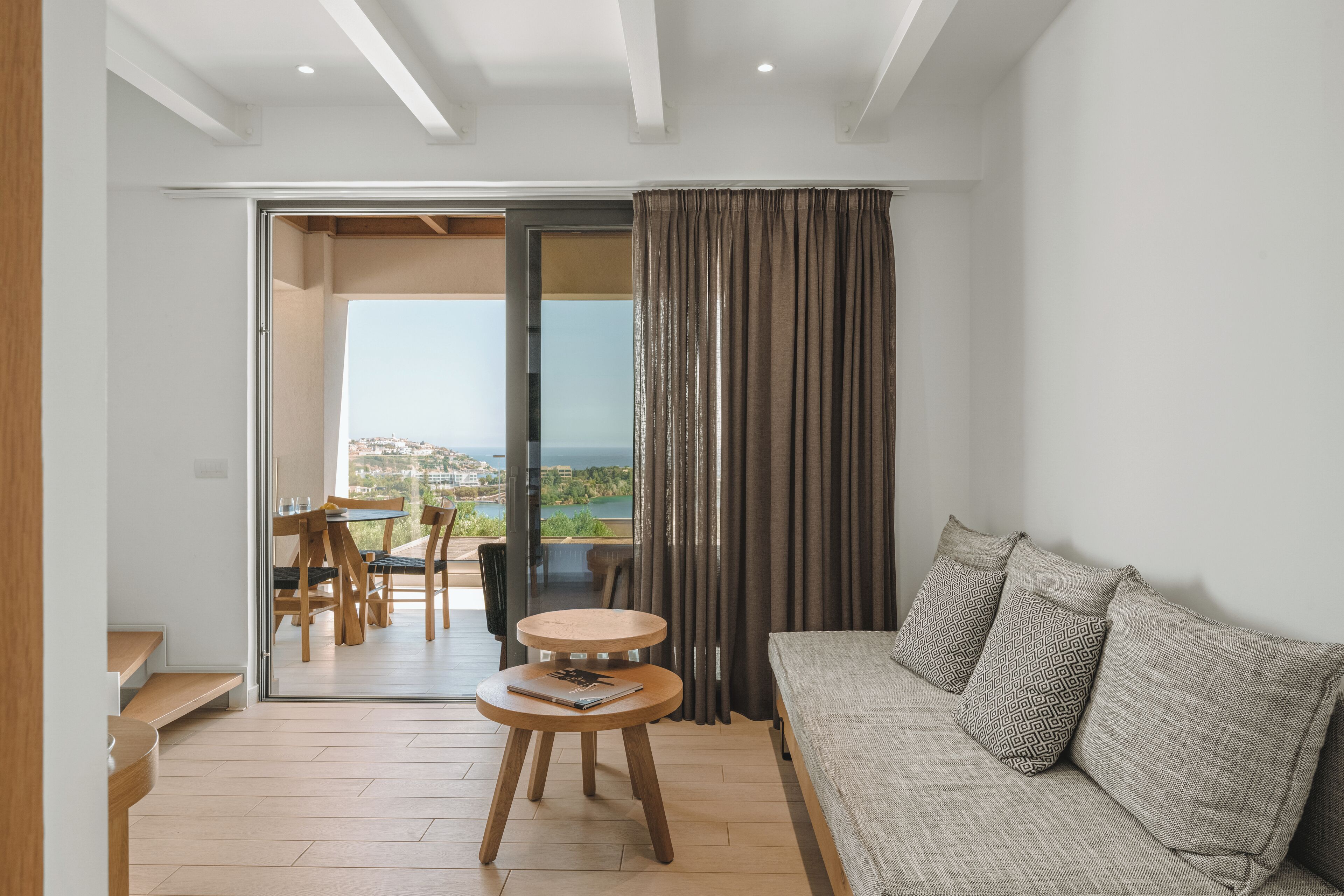 A modern living room with a sofa, a small round table with a book, and a view of the outdoors through a glass door.