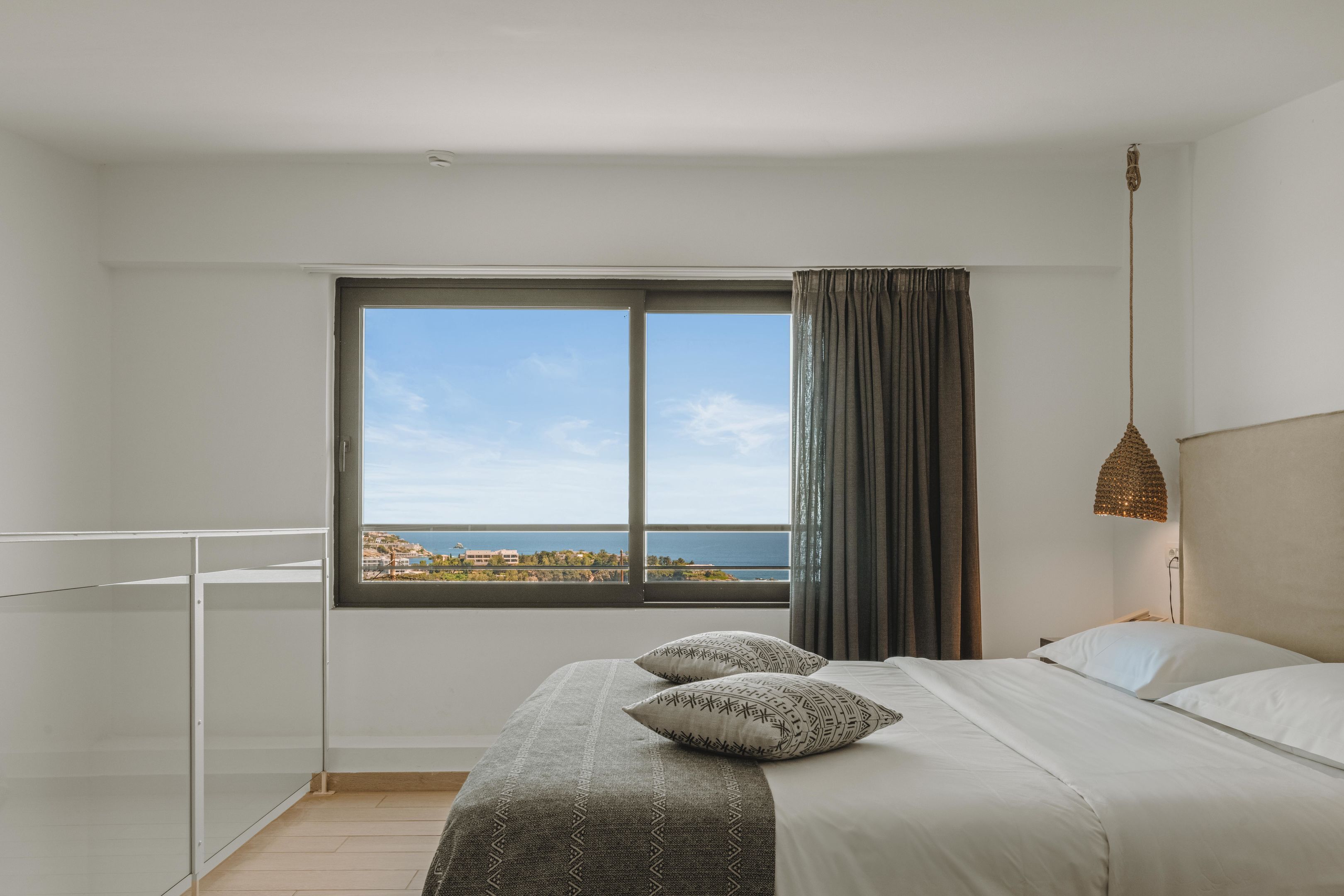 A modern bedroom with a large window, a bed with patterned pillows, and a view of the sea.