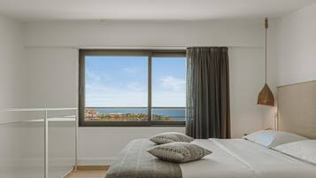 A modern bedroom with a large window, a bed with patterned pillows, and a view of the sea.