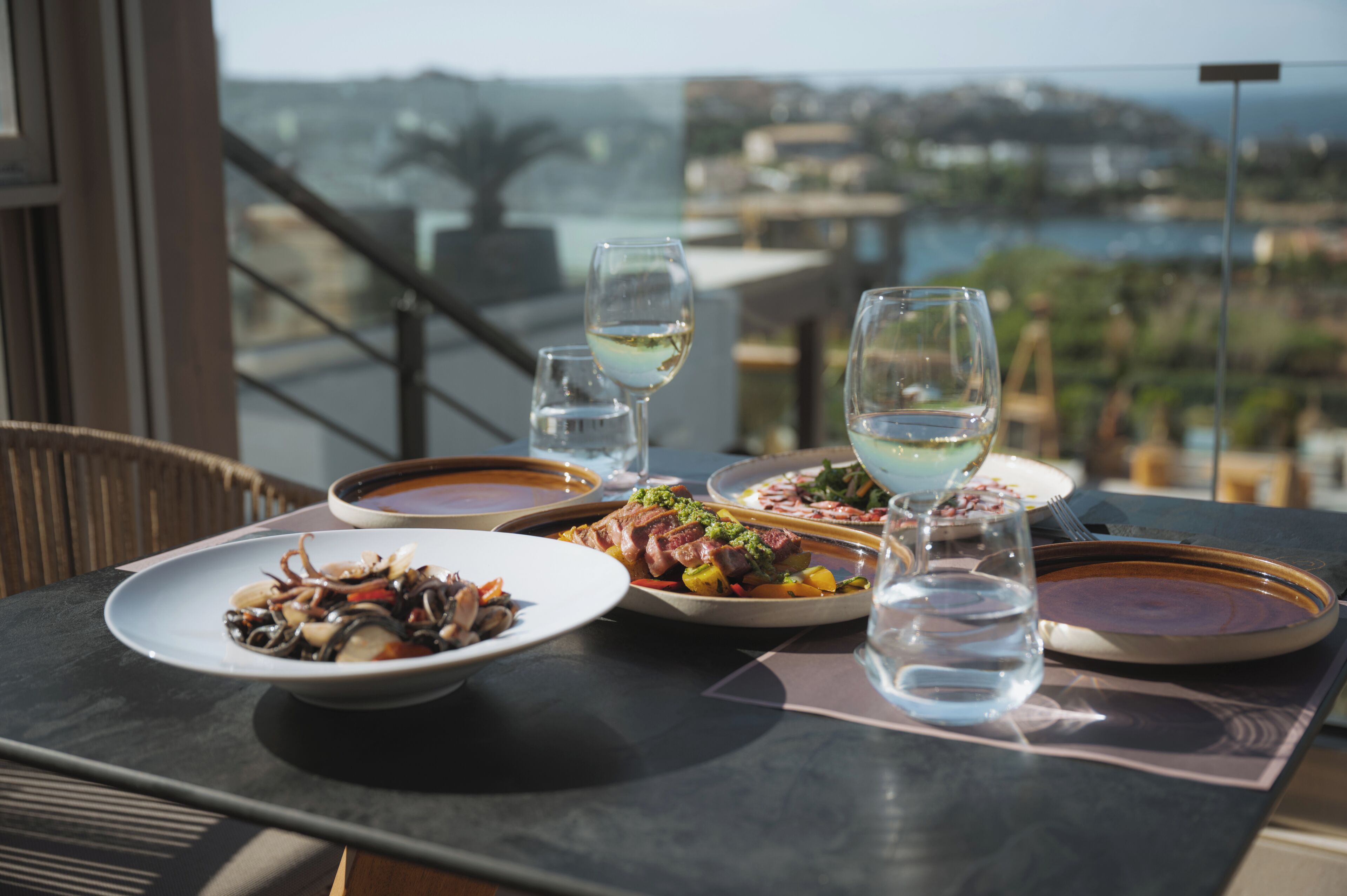 A table set with various dishes and glasses of water, overlooking a scenic view of a coastal area.