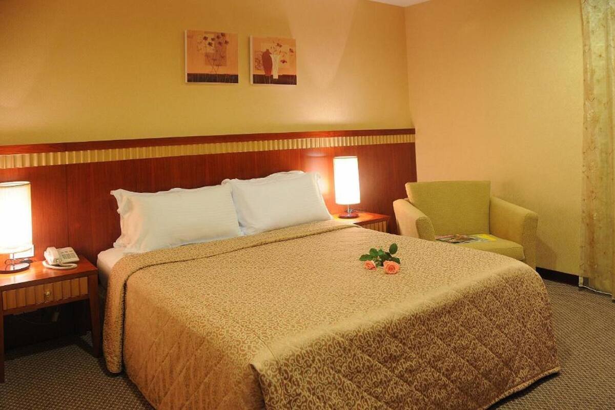A hotel room with a large bed, two bedside lamps, a nightstand with a phone, a small table with a magazine, and a chair.
