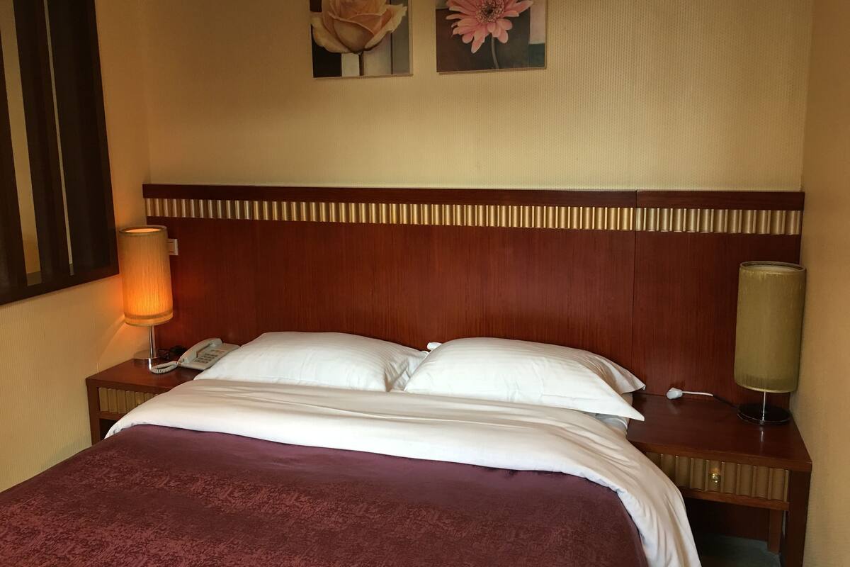 A hotel room with a wooden headboard, a bed with white linens, a red throw blanket, two bedside lamps, and two wall-mounted flower paintings.