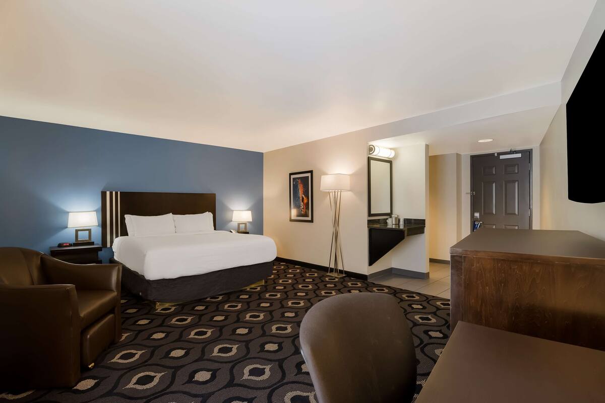 A hotel room with a bed, two armchairs, a desk, a lamp, a mirror, and a patterned carpet.