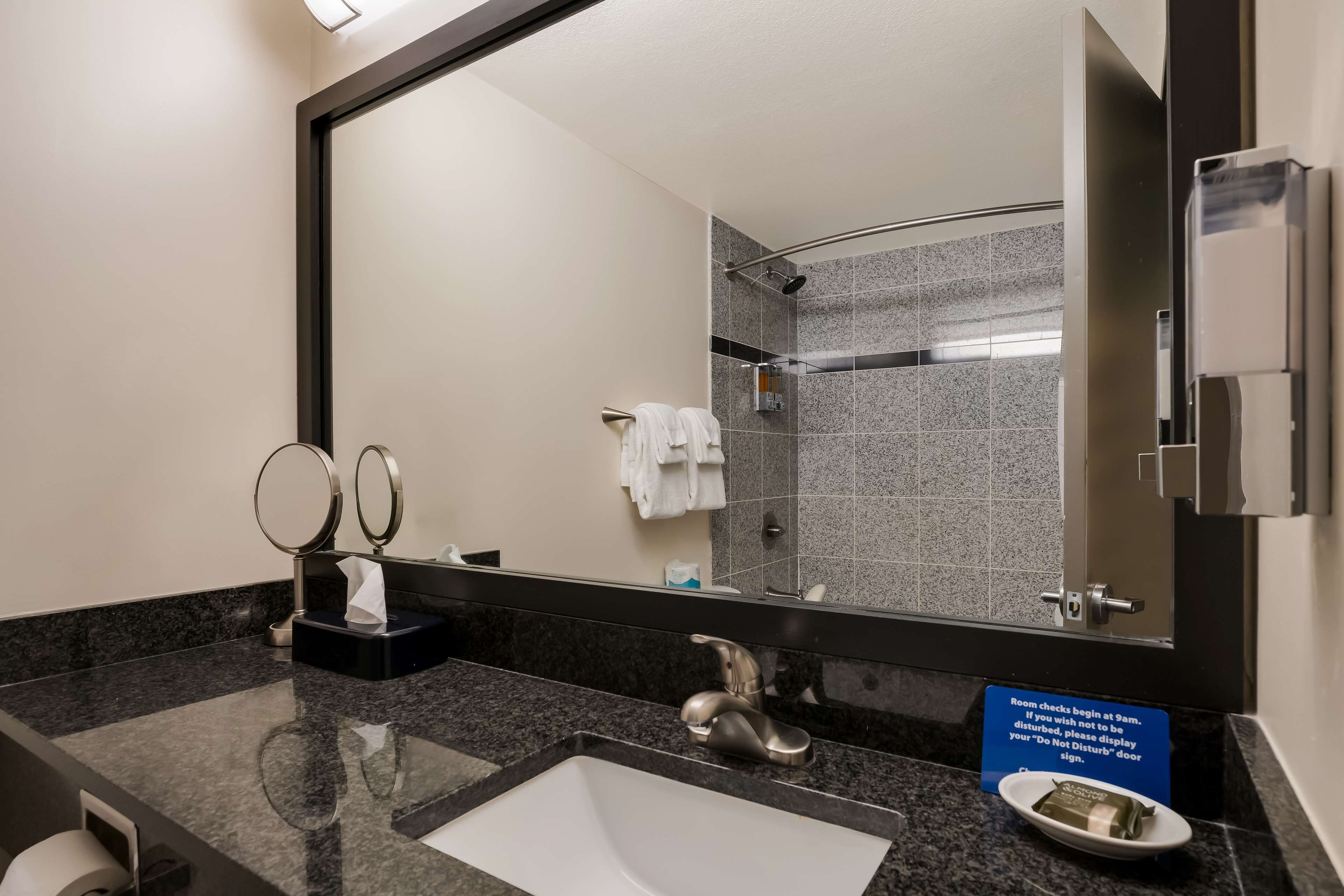 A hotel bathroom with a large mirror, a sink, a towel rack, and a sign indicating room check-in times.