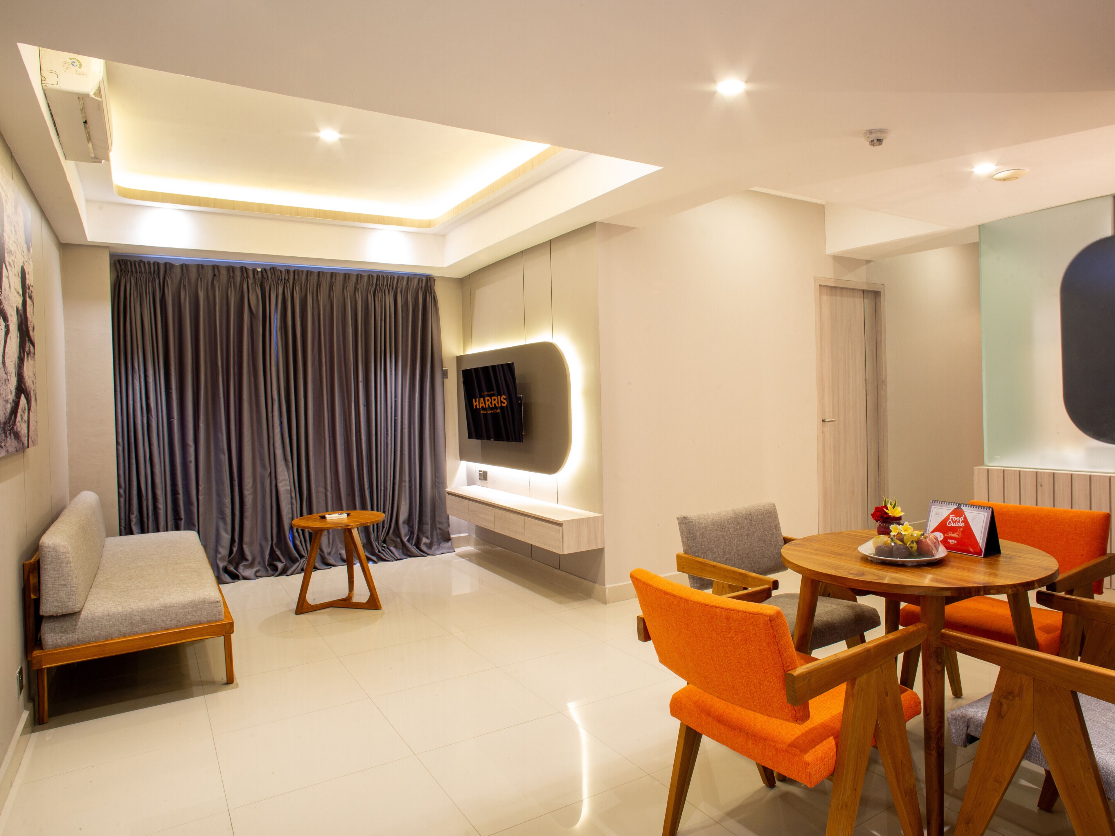 A modern hotel room with a sofa, a dining table with chairs, a TV mounted on the wall, and a large window with curtains.