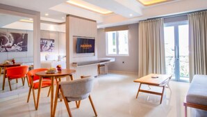 Een moderne hotelkamer met een flatscreen-tv, een zithoek met oranje stoelen, een grijze bank en een balkon met gordijnen.