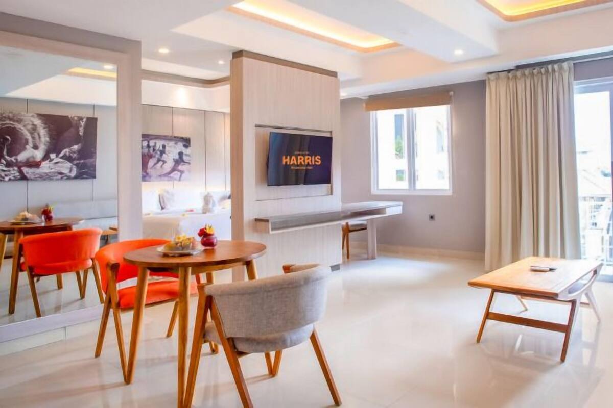 Ein modernes Hotelzimmer mit Flachbildfernseher, einer Essecke mit orangefarbenen Stühlen, einem grauen Sofa und einem Balkon mit Vorhängen.