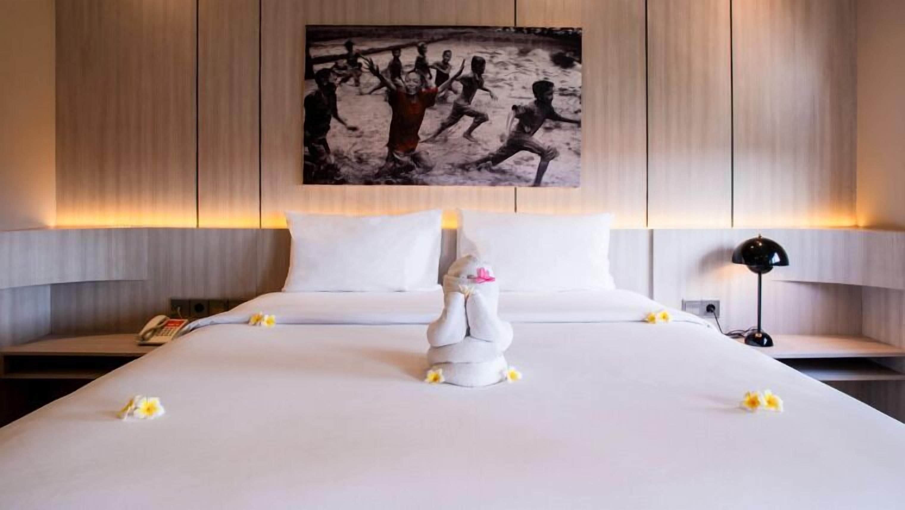 A hotel room with a large bed, white linens, a decorative pillow, and a painting on the wall.