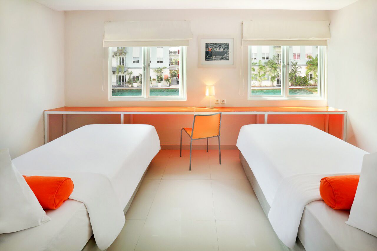 A hotel room with two beds, a desk, a chair, and large windows with a view of buildings and palm trees.