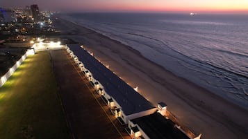 A beachfront hotel with a row of rooms along the shore, a grassy area, and a view of the ocean.