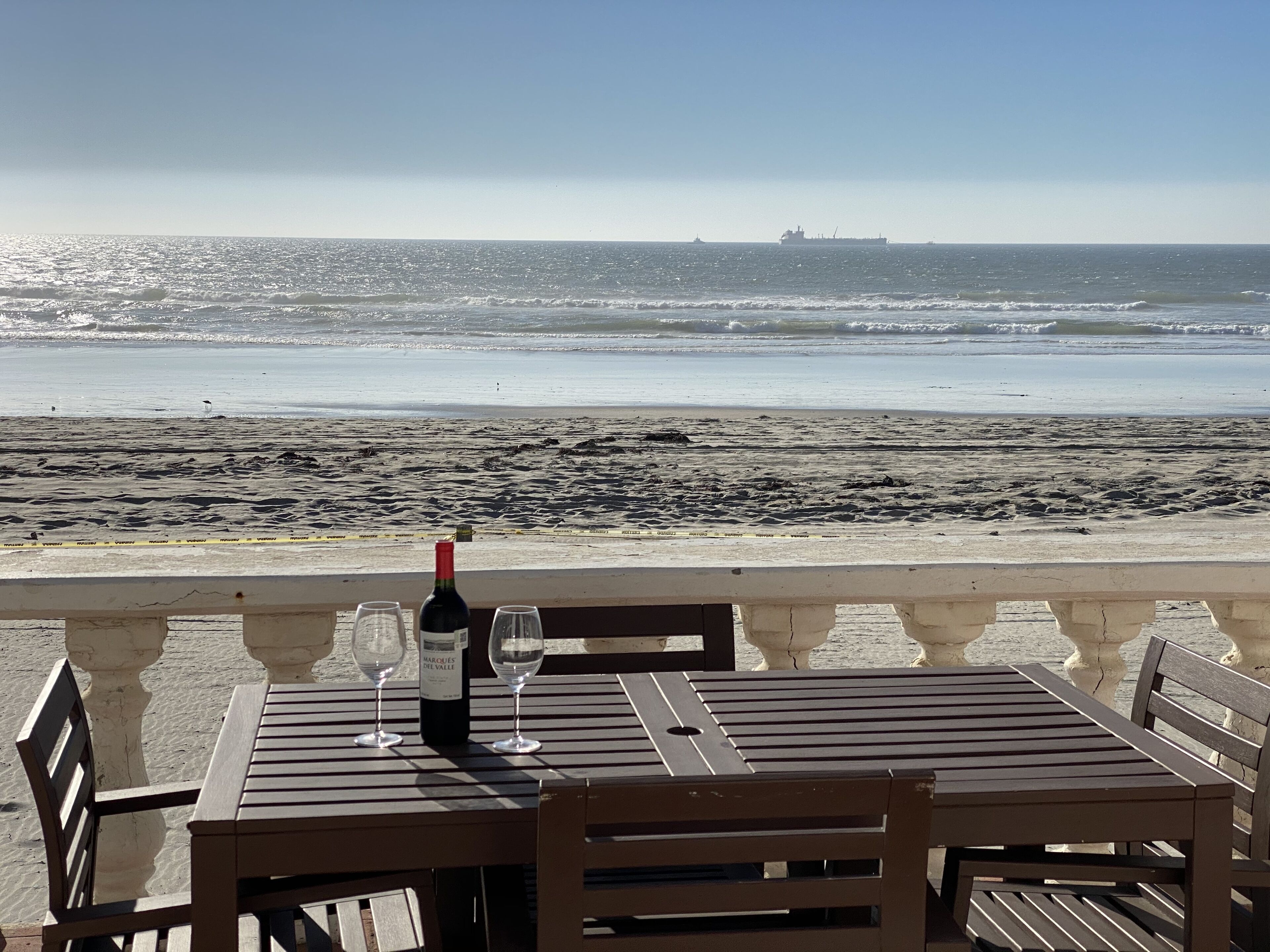 A table with a bottle of wine and two glasses, overlooking a beach with a ship in the distance.