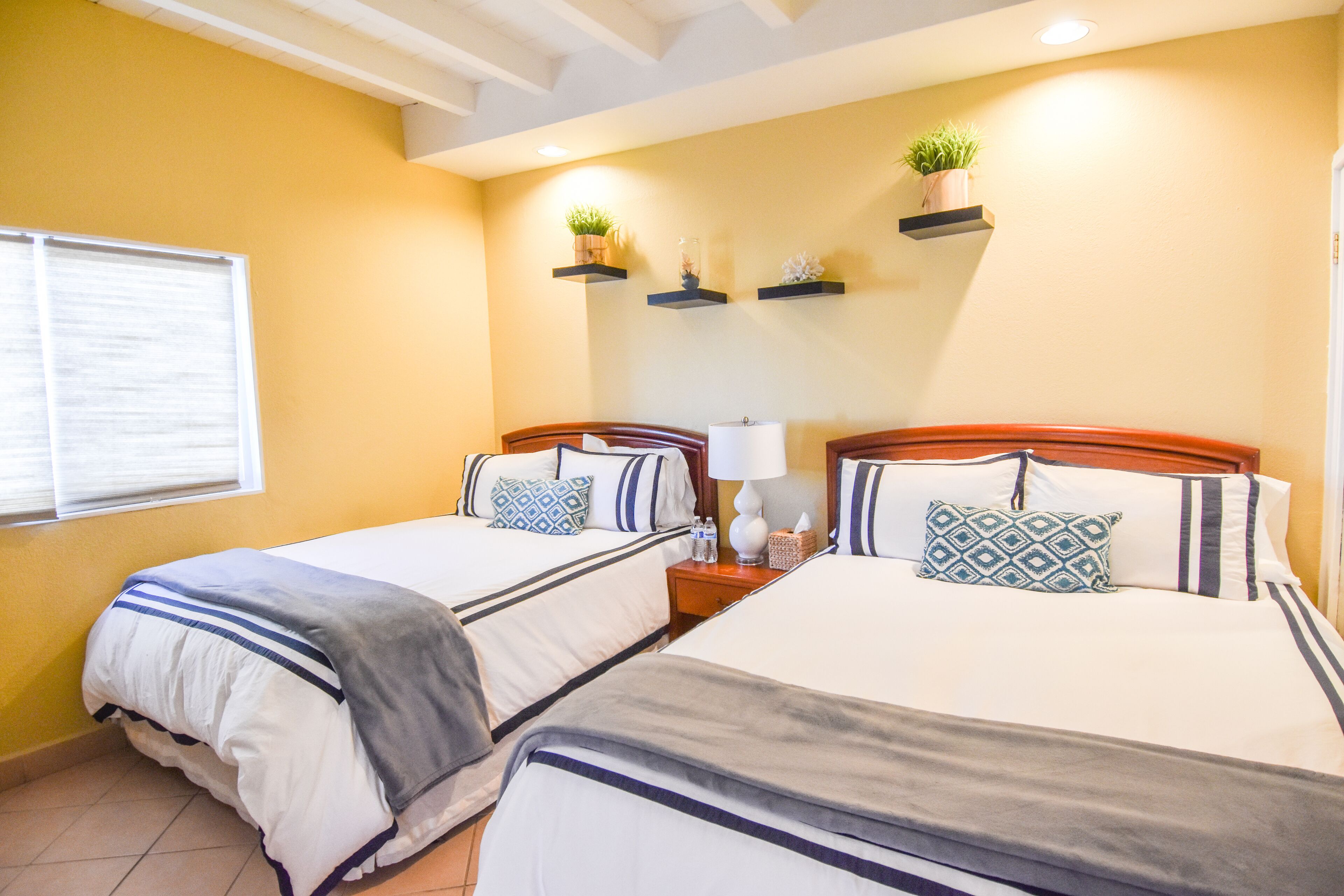 Habitación de hotel con dos camas, cada una con cabecera, mesitas de noche y almohadas decorativas. Hay estantes en la pared con plantas y una ventana con persianas.