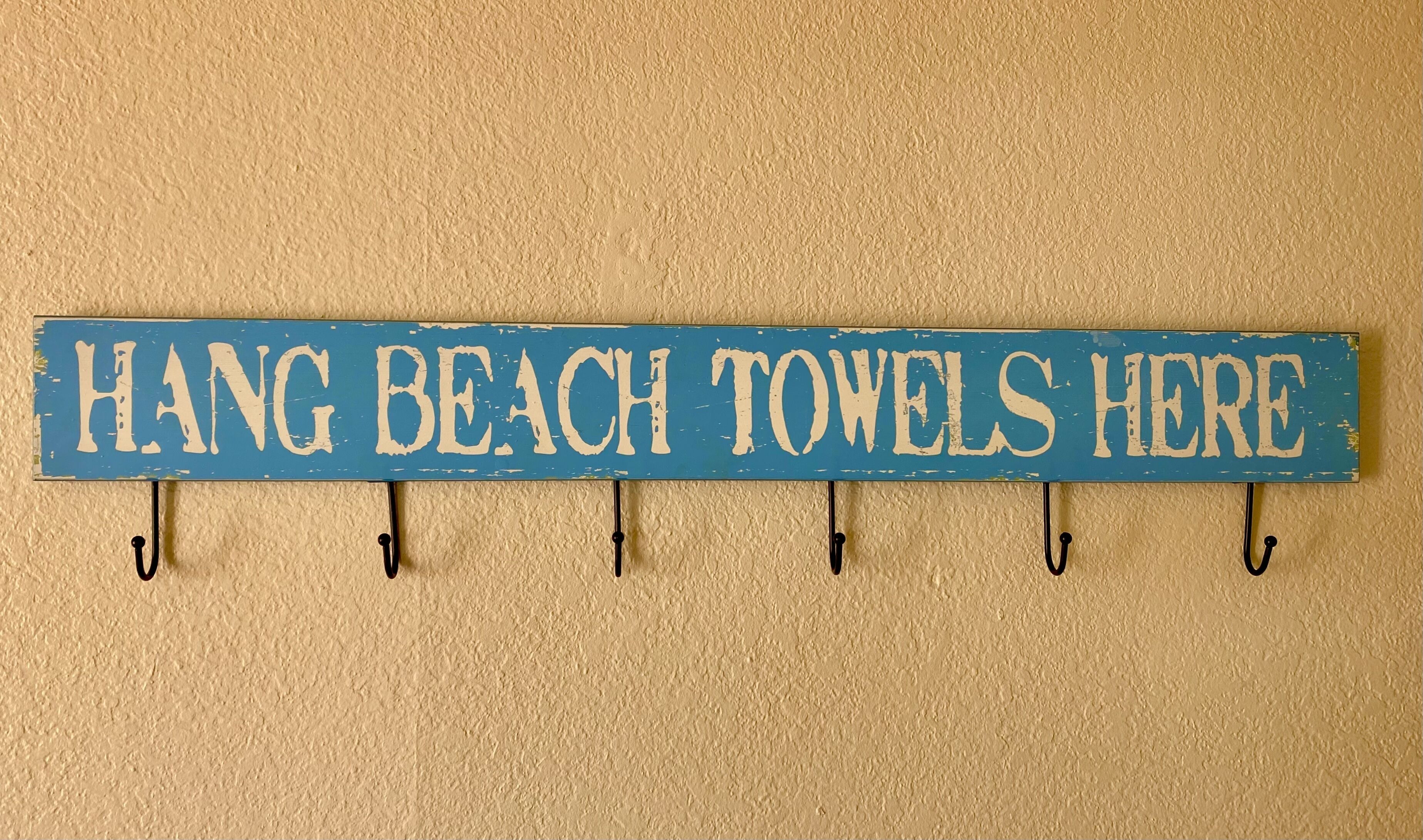 A blue sign with white text that reads "HANG BEACH TOWELS HERE" mounted on a wall with hooks.