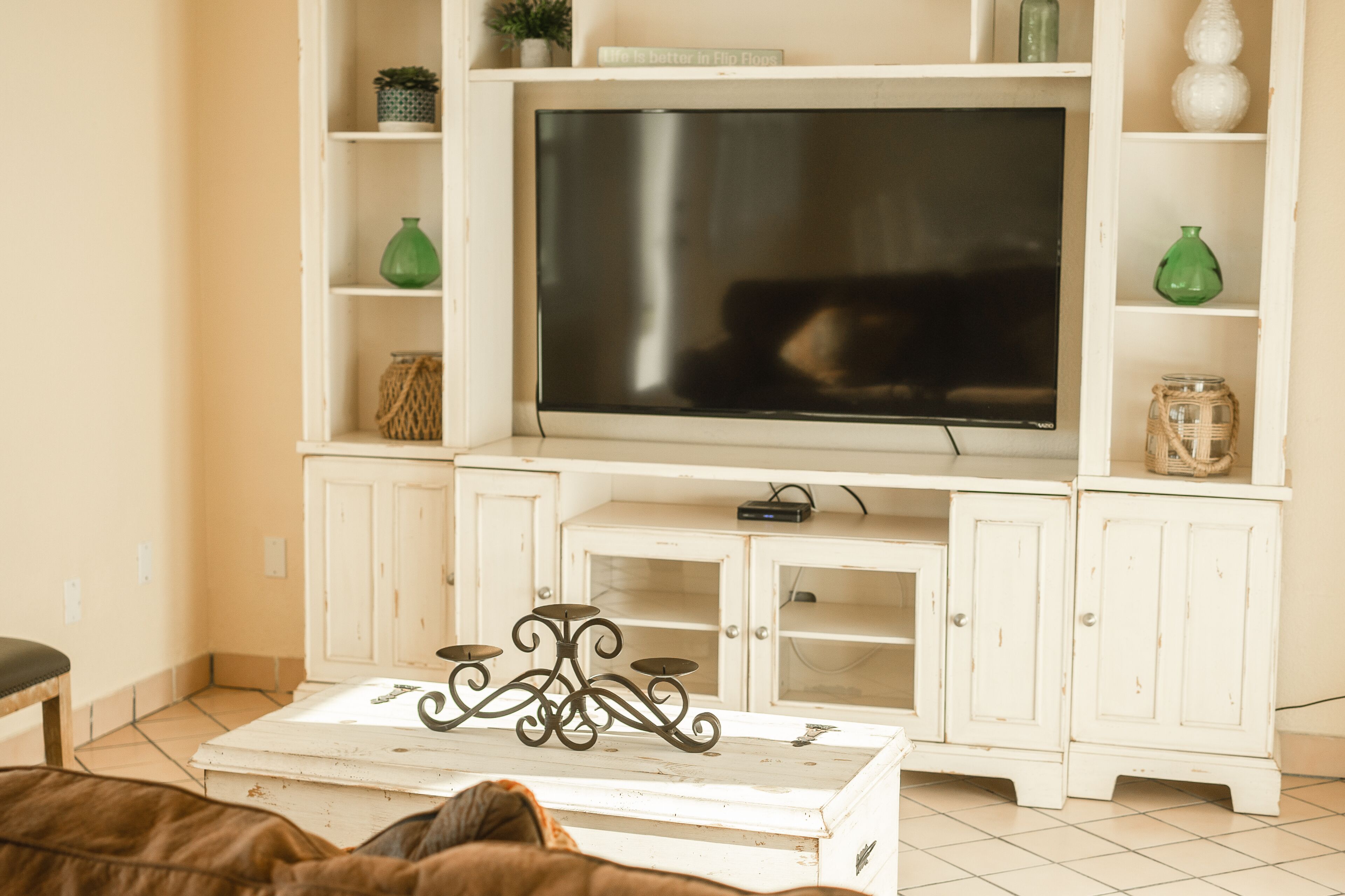 Una sala de estar con un televisor de pantalla plana grande sobre un mueble blanco, una mesa de centro con un soporte metálico decorativo y estanterías con varios objetos, incluyendo jarrones verdes.