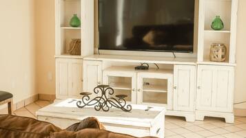 Una sala de estar con un televisor de pantalla plana grande sobre un mueble blanco, una mesa de centro con un soporte metálico decorativo y estanterías con varios objetos, incluyendo jarrones verdes.
