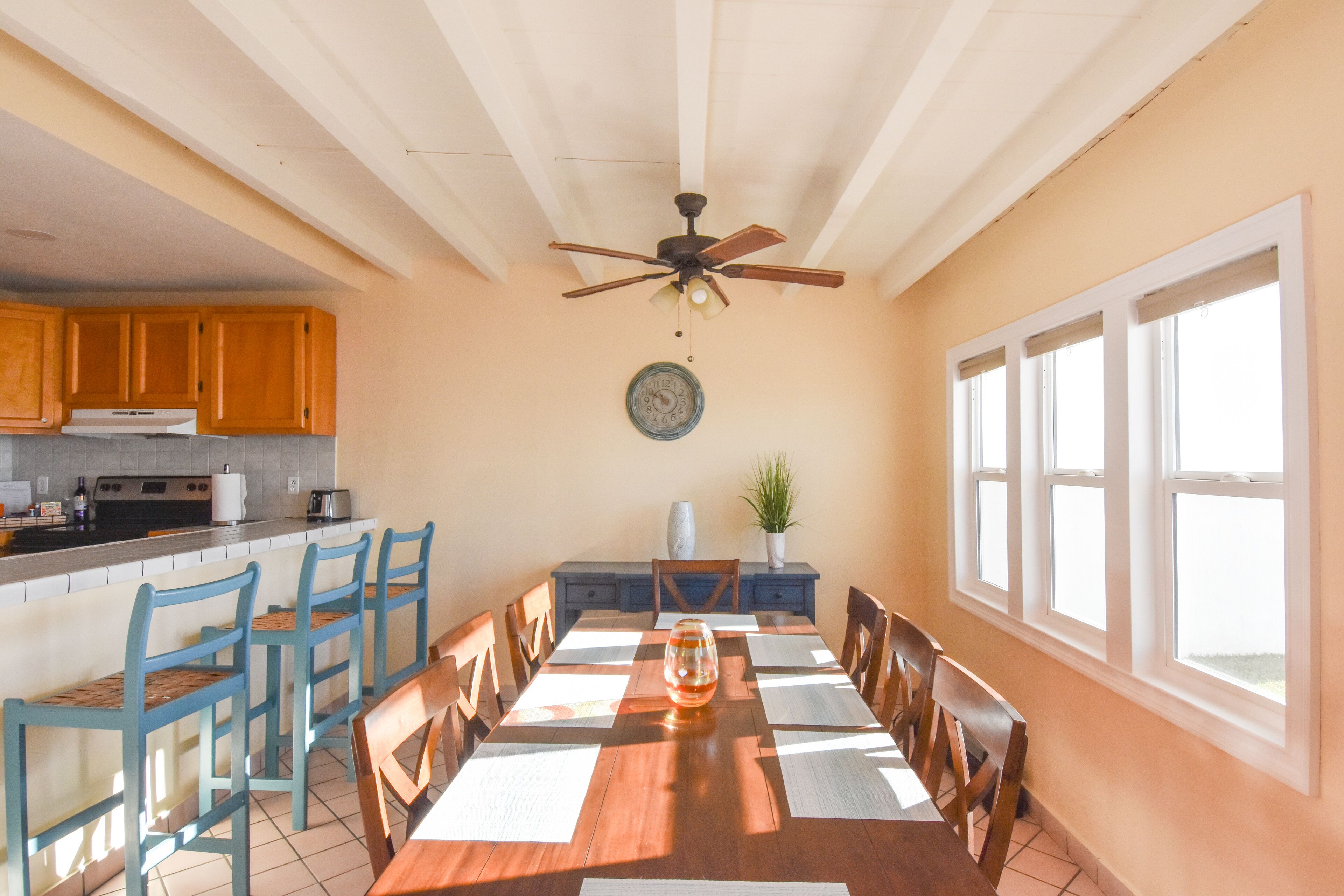A dining area with a wooden table and chairs, a ceiling fan, a clock, and a window with blinds.