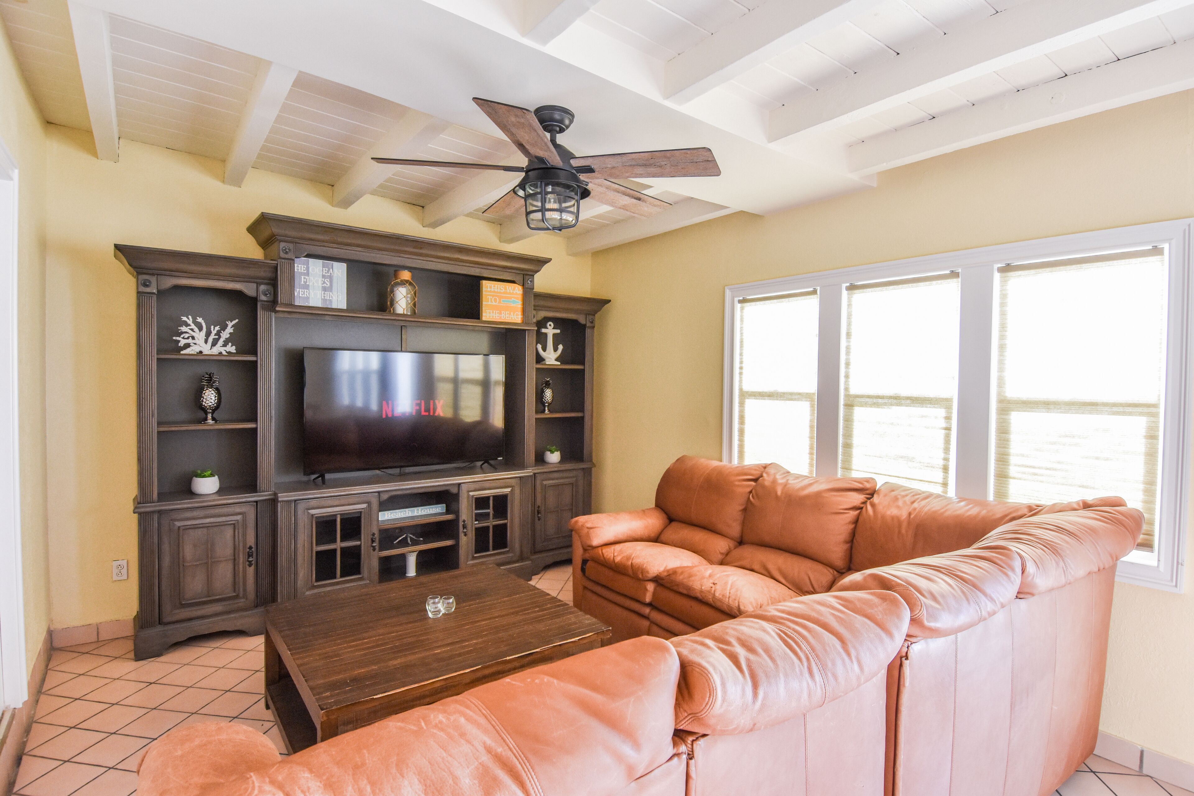 A living room with a leather sofa, a wooden coffee table, a TV on a wooden stand, and a ceiling fan.