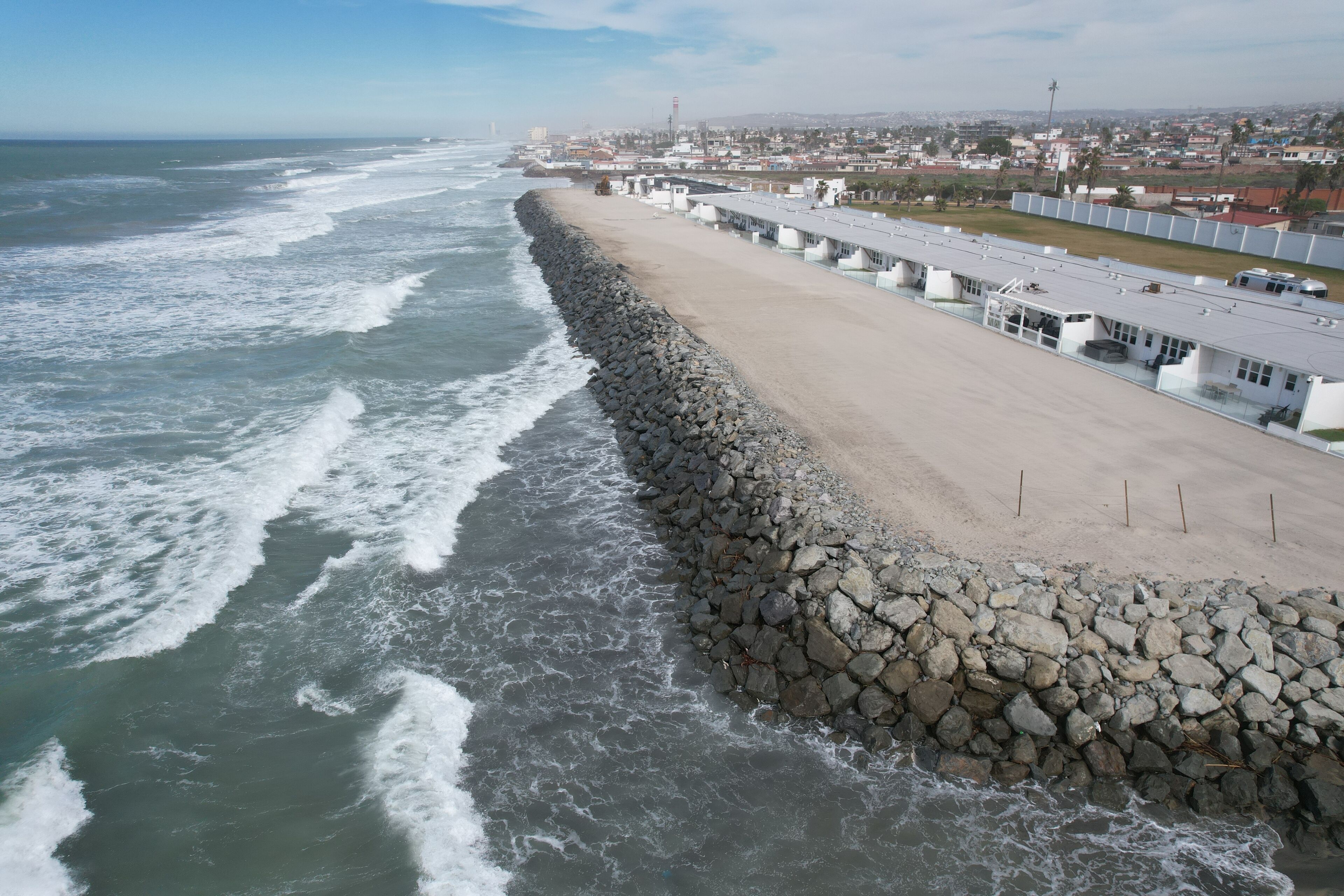 A coastal view with a rocky breakwater, a sandy beach, and a row of buildings with a flat roof.