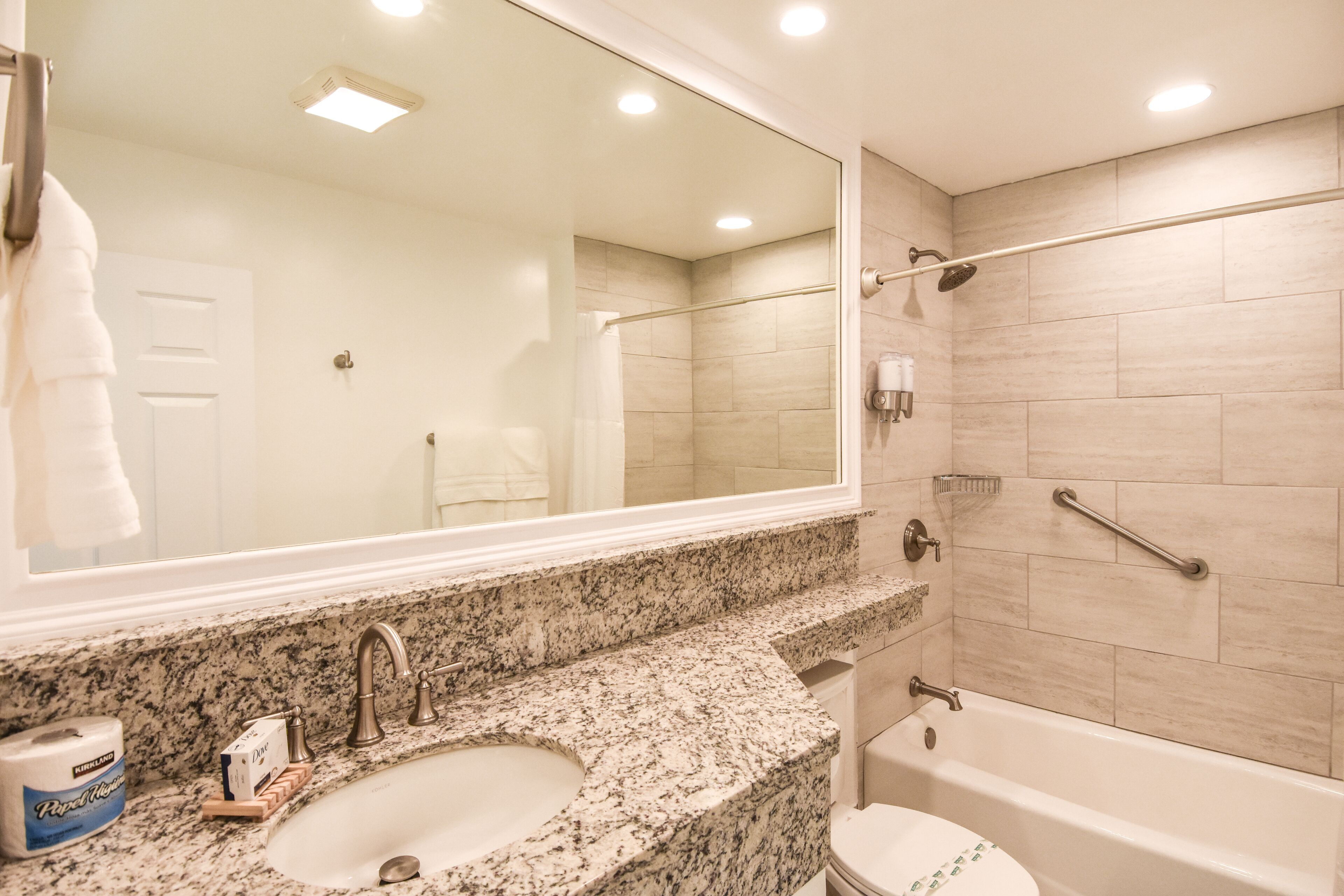 Baño con lavabo de encimera de granito, un espejo grande, bañera y zona de ducha.