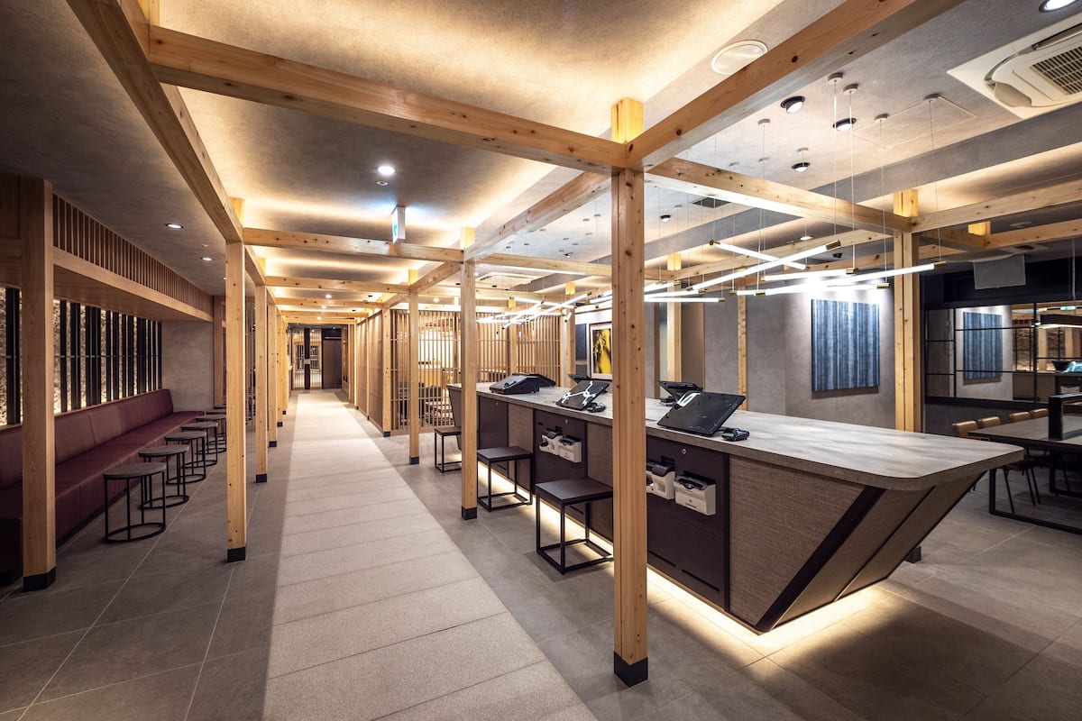 Un interior moderno con un largo pasillo, vigas de madera y una zona central con mostrador, asientos y mesas.