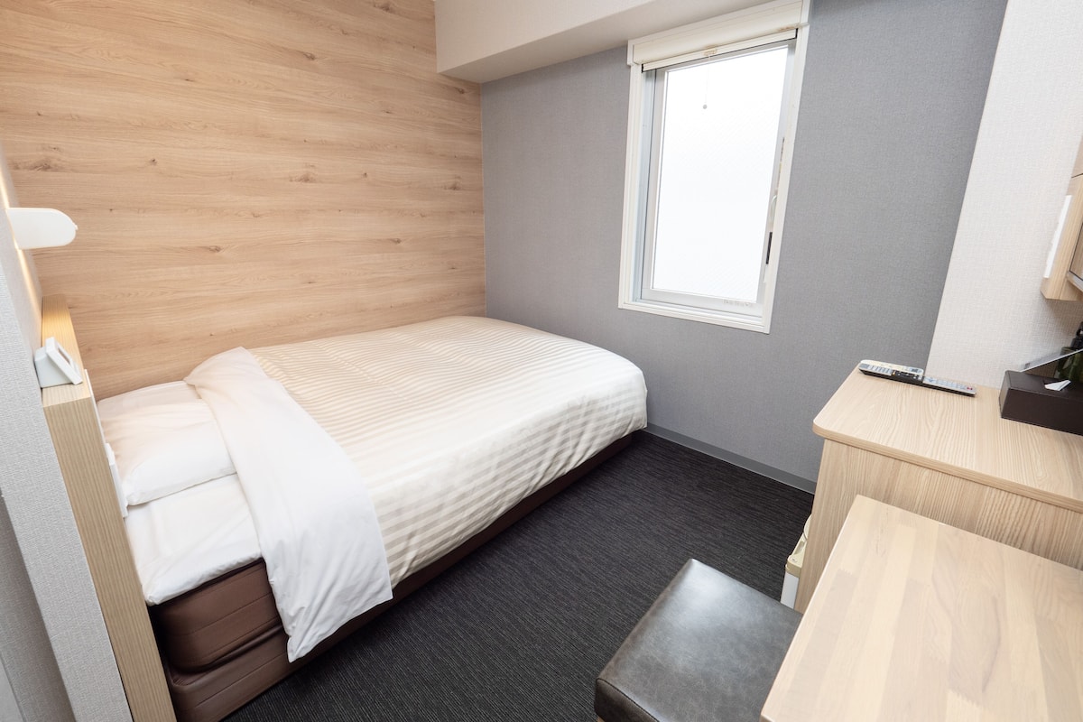 Habitación de hotel pequeña y compacta con una cama individual, una mesita de noche, un escritorio pequeño y una ventana con persianas.