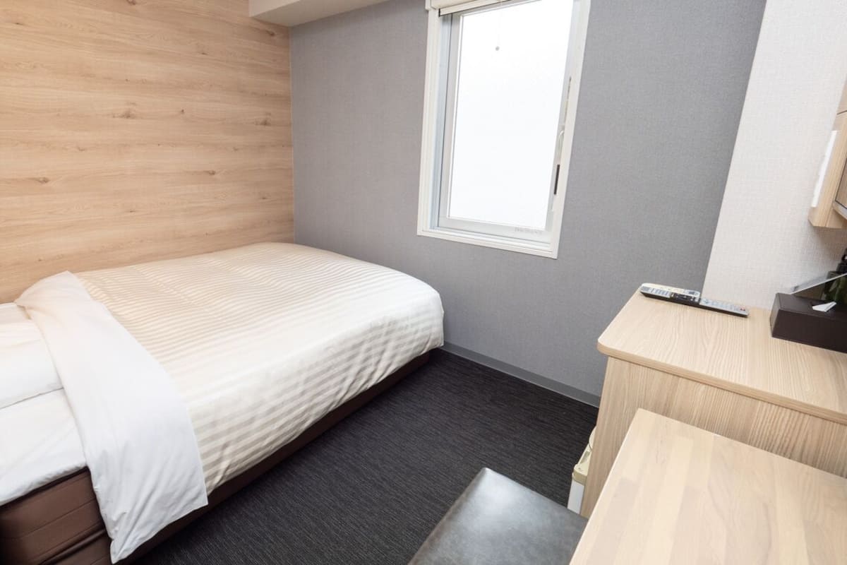 Habitación de hotel compacta con una cama individual, un escritorio pequeño, una ventana y una pared gris.