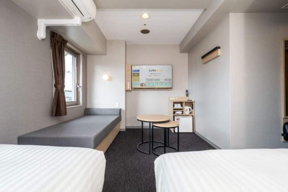 A hotel room with two beds, a small table, a bench, a window with curtains, a wall-mounted screen, and a shelf with items.