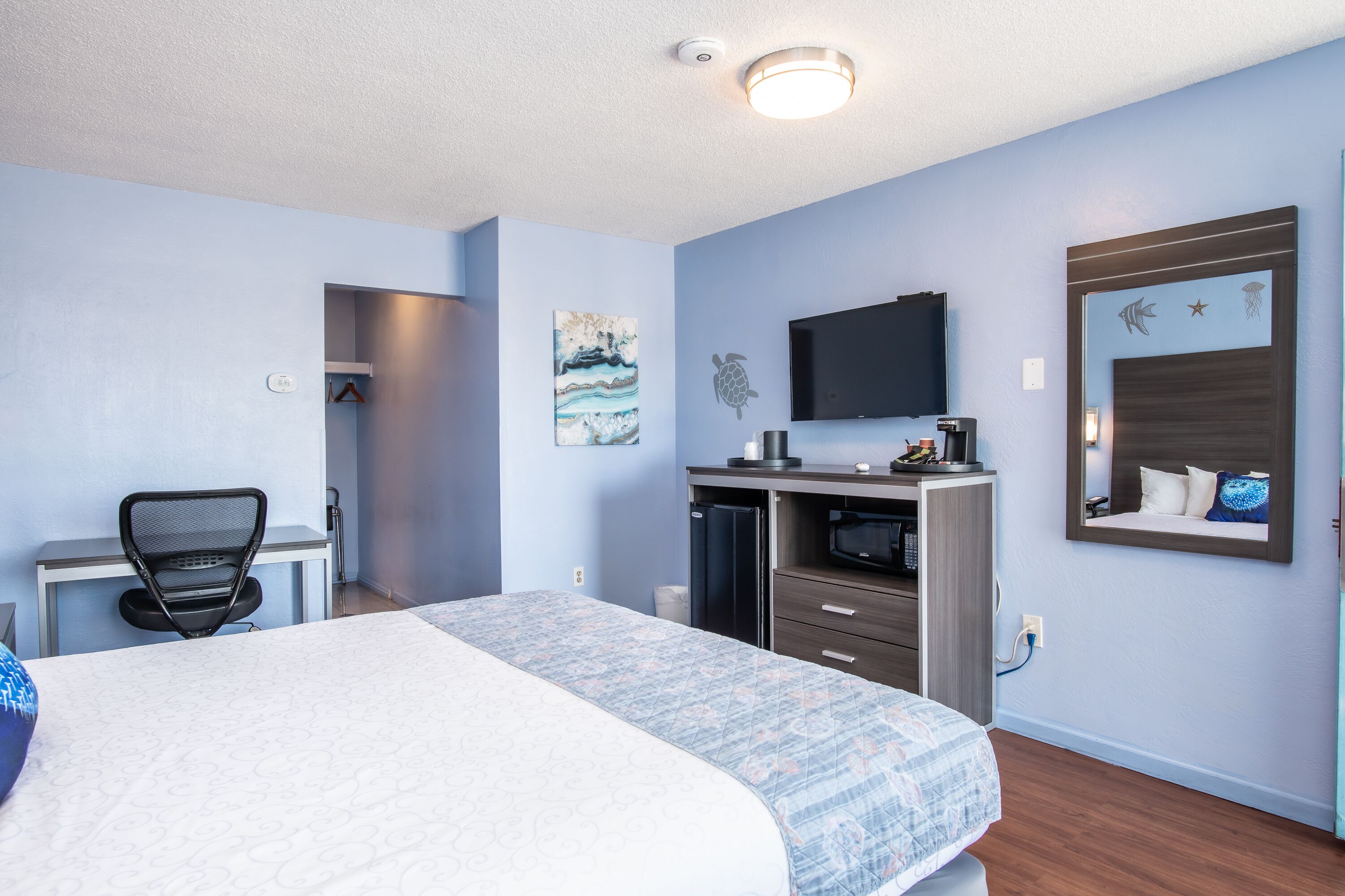 A hotel room with a bed, desk, chair, TV, and a small kitchenette.