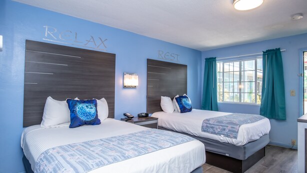 A hotel room with two beds, a nightstand, a lamp, and a window with curtains. - Aqua Breeze Inn (Santa Cruz)