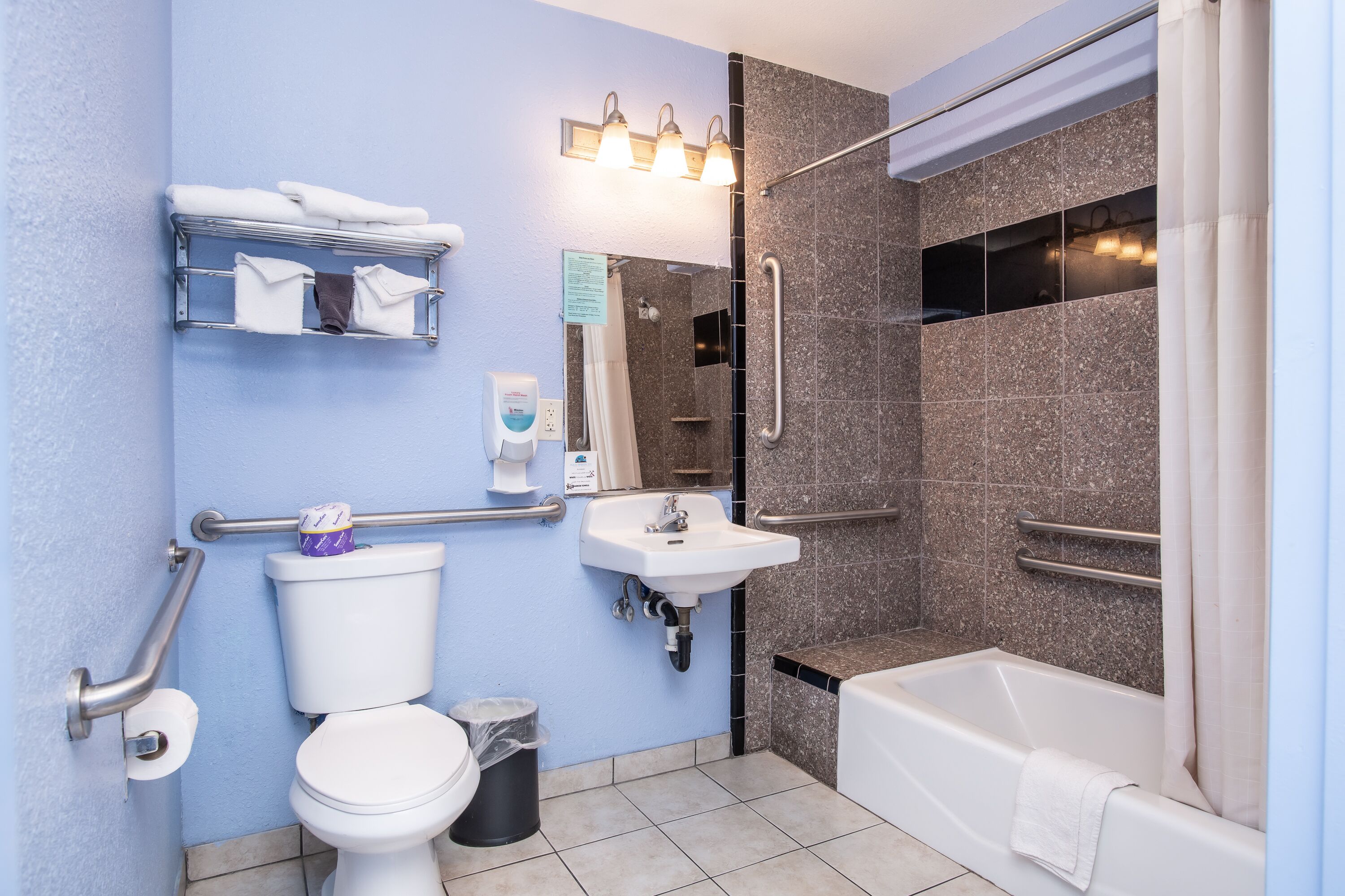 A bathroom with a toilet, sink, and bathtub. There are grab bars, a towel rack, and a trash can.