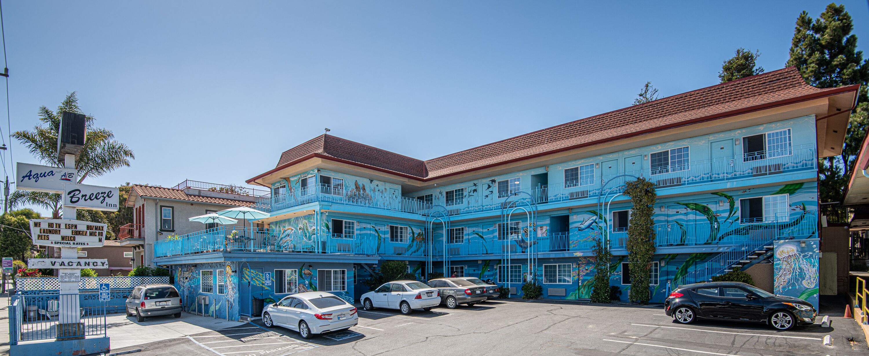 Un immeuble à plusieurs étages, à la façade bleue, orné de peintures murales représentant la vie marine. Plusieurs voitures sont garées devant, et on peut voir une enseigne indiquant « Breeze Inn ».