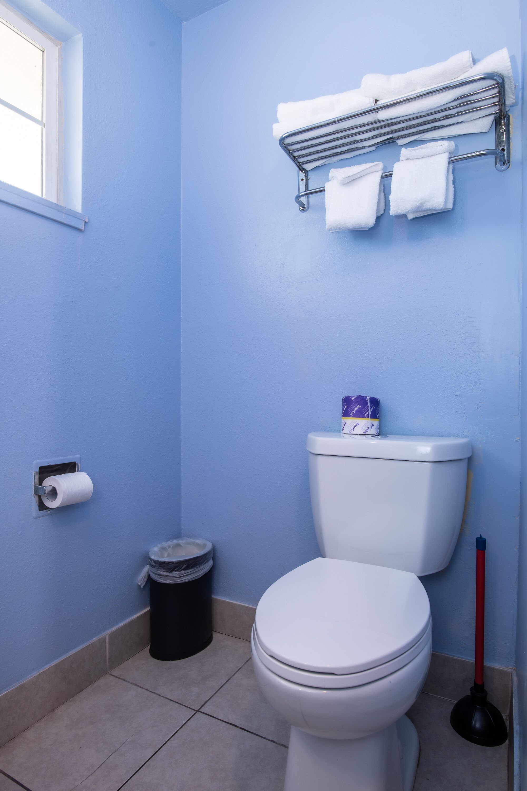Ein Badezimmer mit Toilette, Mülleimer, Toilettenpapierhalter, Handtuchhalter mit Handtüchern und einem Fenster.
