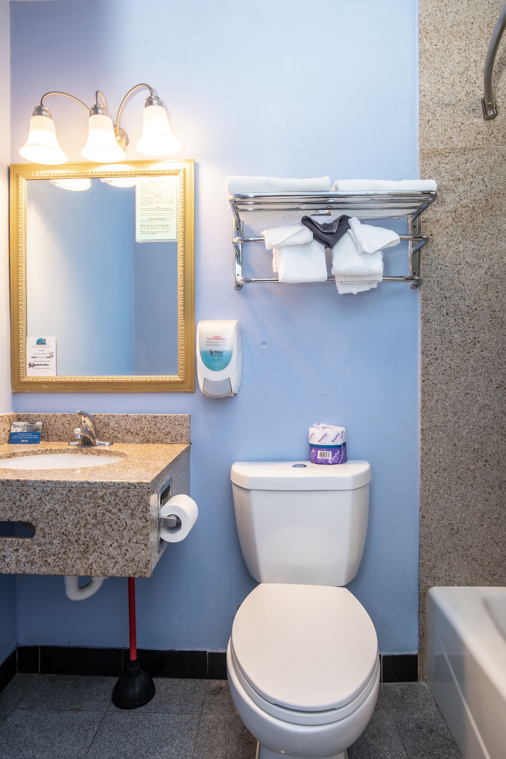 A bathroom with a granite sink, a white toilet, a towel rack with towels, a soap dispenser, and a mirror with lights.
