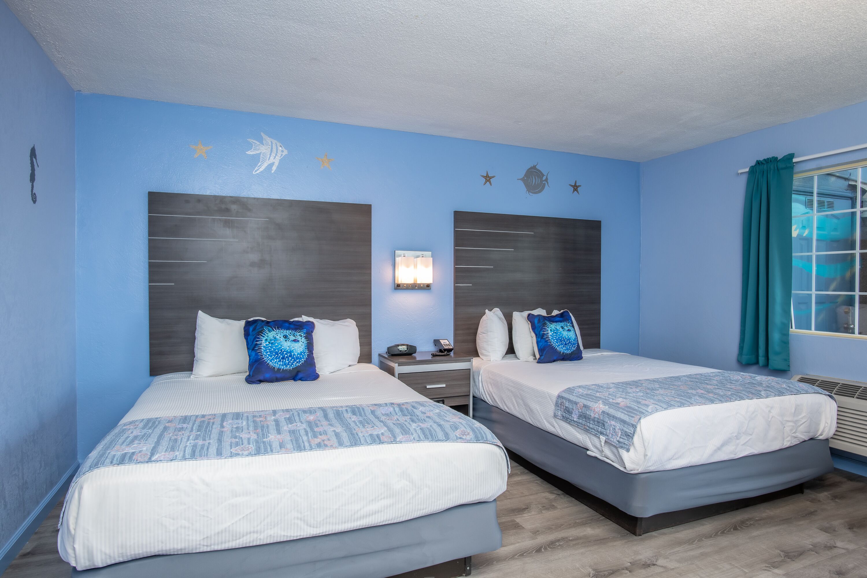 A hotel room with two beds, a nightstand, a wall-mounted lamp, and a window with curtains.