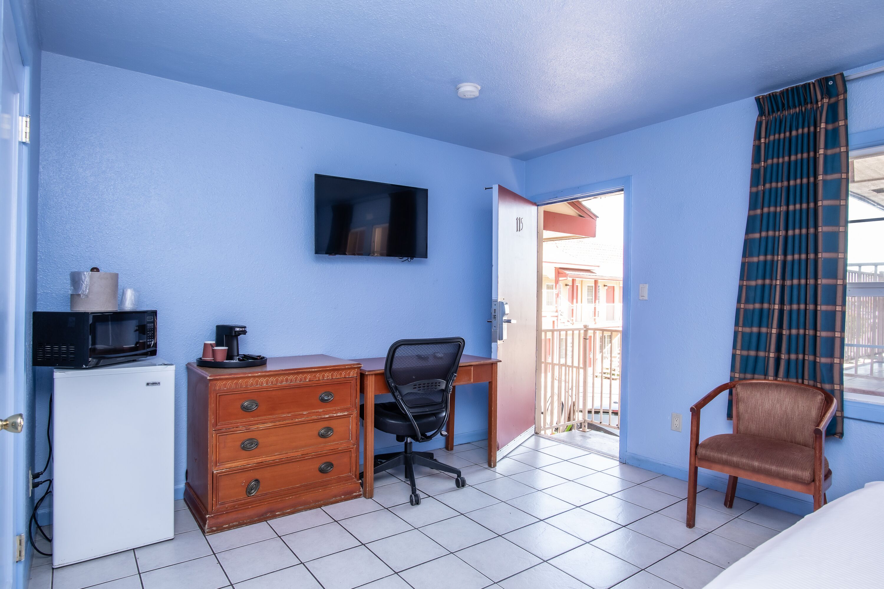 A compact room with a desk, a chair, a small refrigerator, and a television mounted on the wall.