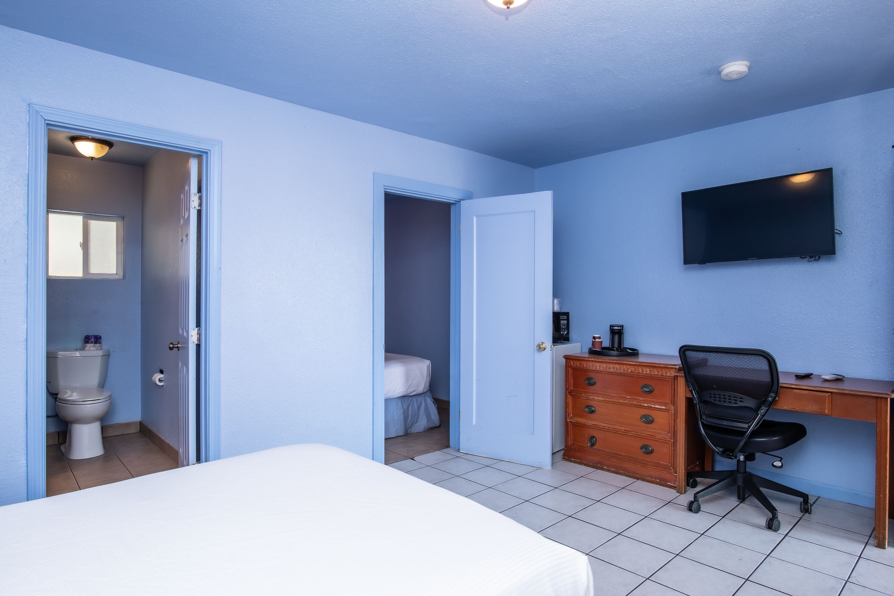 A bedroom with a bed, a desk, a chair, a dresser, a television, and a bathroom with a toilet and sink.