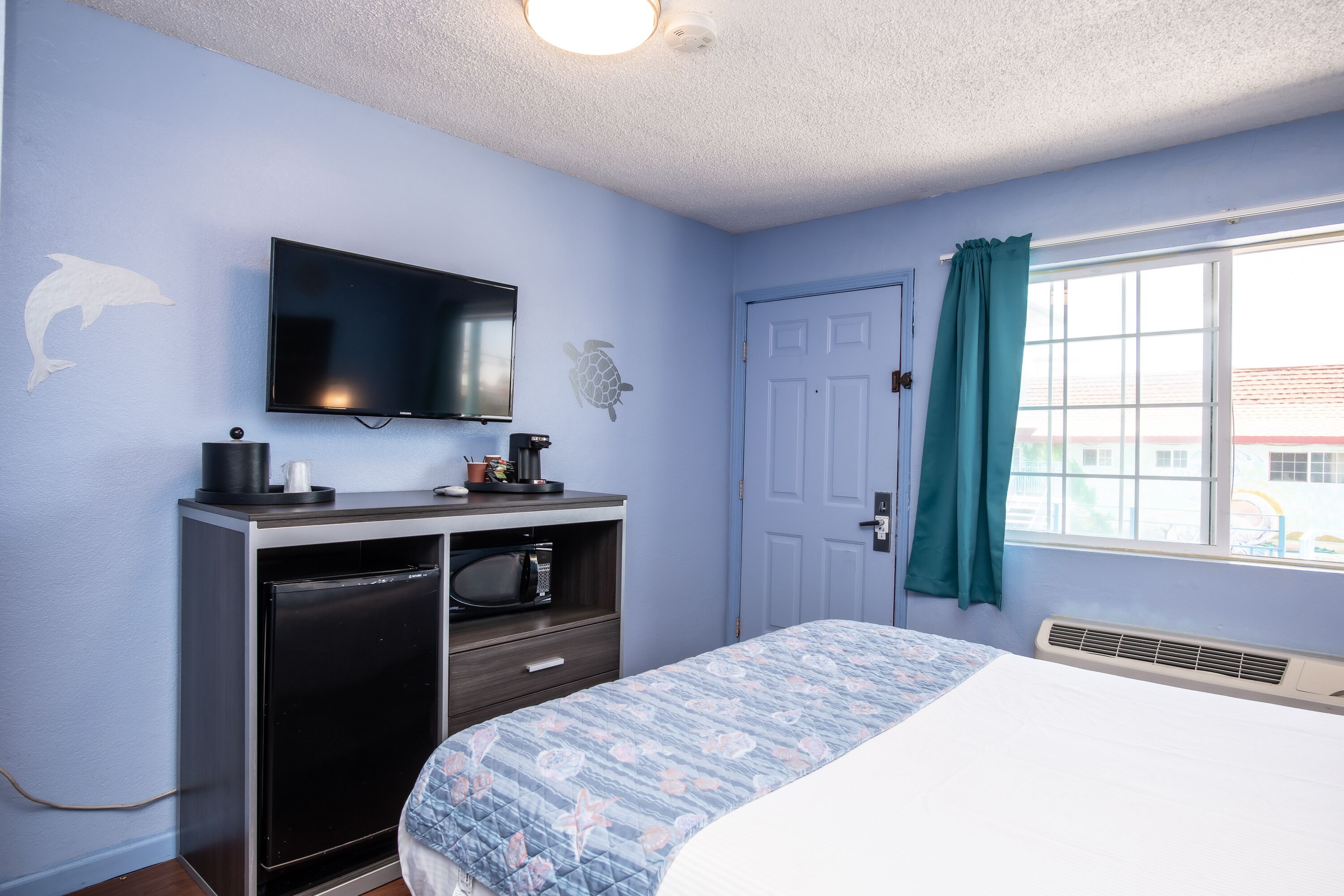 A bedroom with a bed, a TV mounted on the wall, a desk with a microwave, and a door with a curtain.