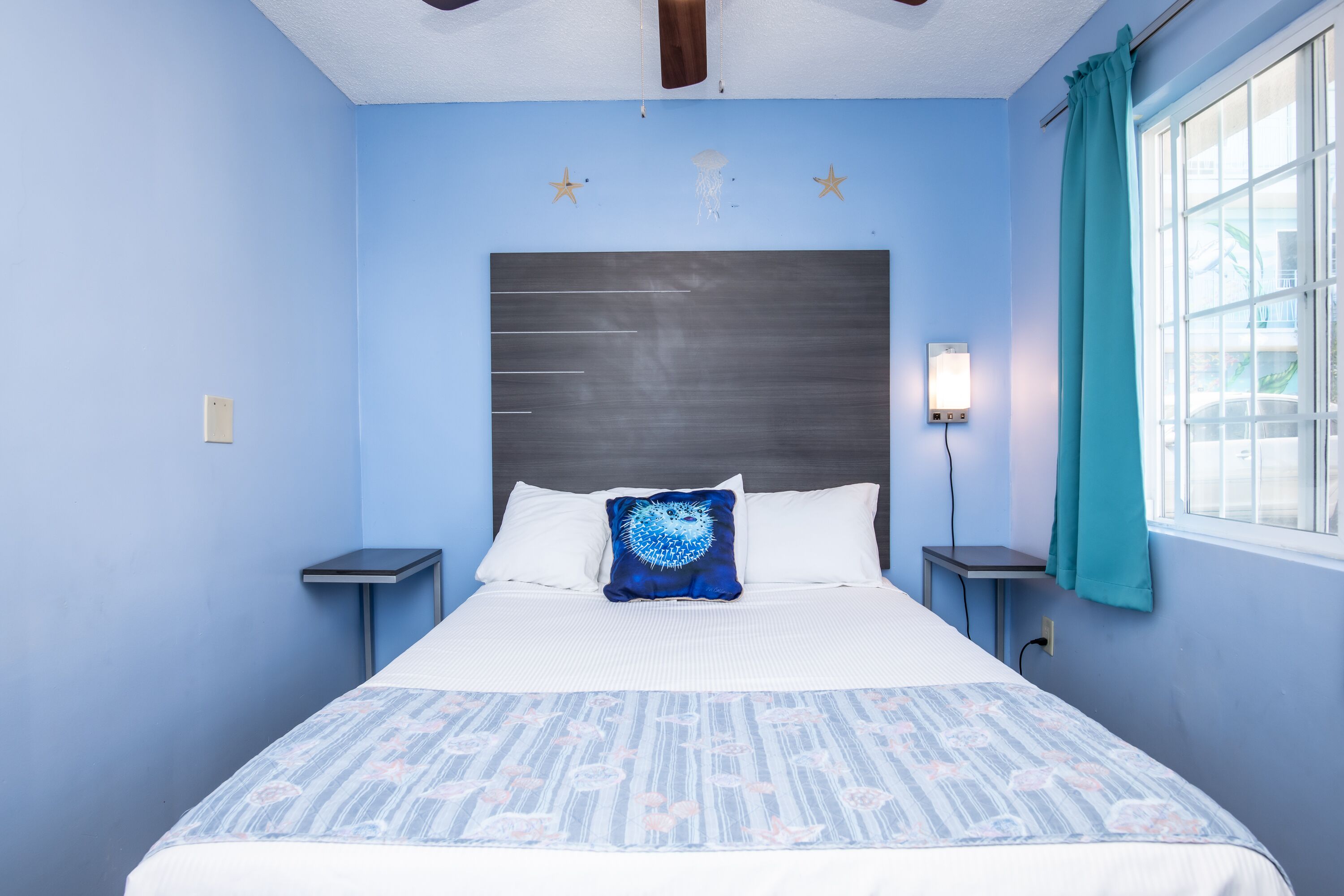 Un dormitorio con cama, mesita de noche, lámpara de pared y ventana con cortinas.