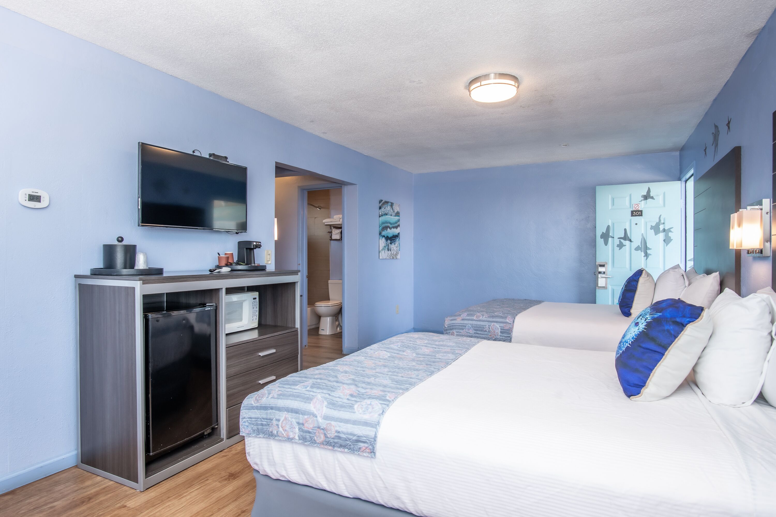 A hotel room with a bed, a desk with a refrigerator, a television, and a bathroom visible through an open door.