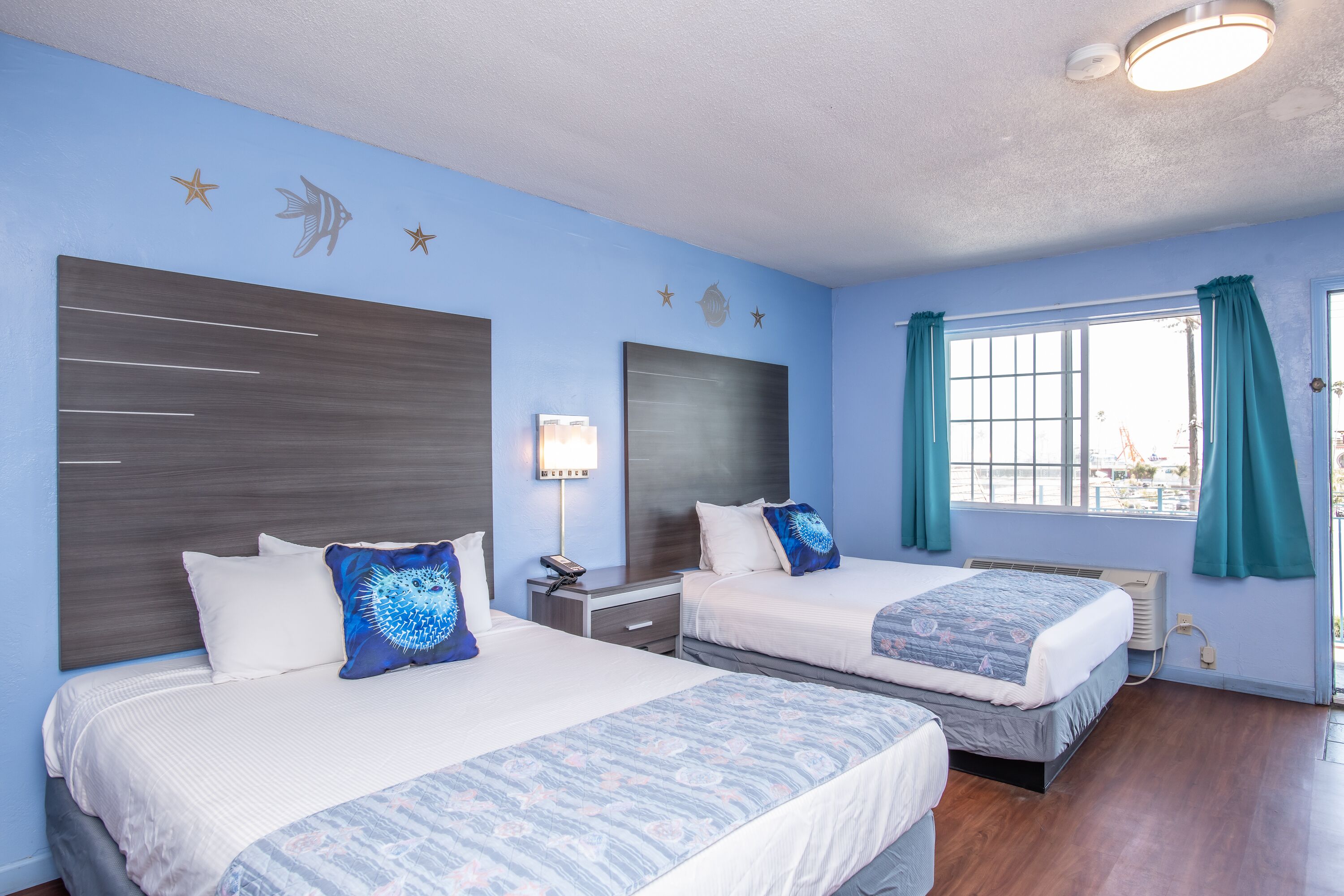 A hotel room with two beds, a nightstand, a lamp, a window with curtains, and a view of the outside.
