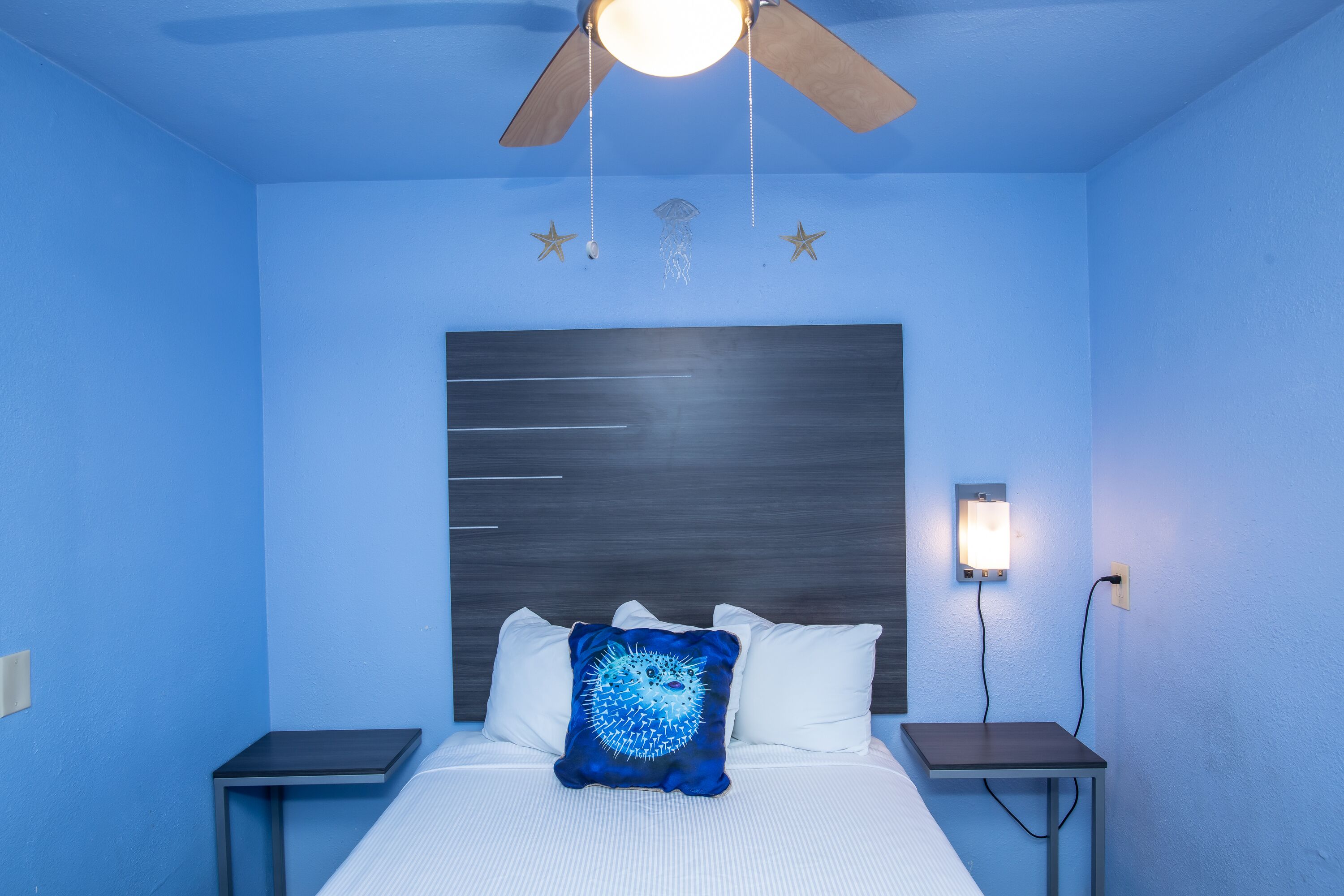 A bedroom with a bed, a ceiling fan, a nightstand, and a wall-mounted light.