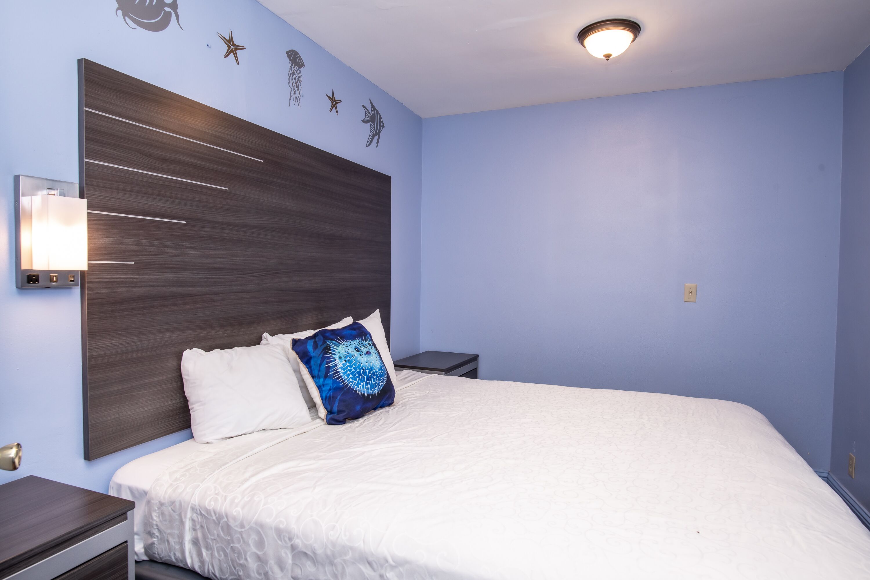 Une chambre à coucher avec un grand lit, une table de chevet, une lampe fixée au mur et des œuvres d’art décoratives accrochées au mur.