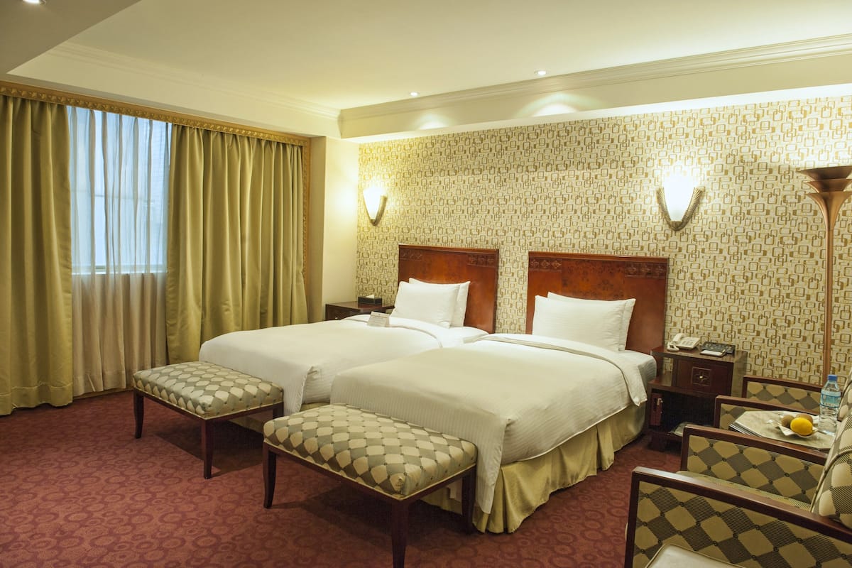 A hotel room with two beds, a bench, a nightstand, and a lamp.