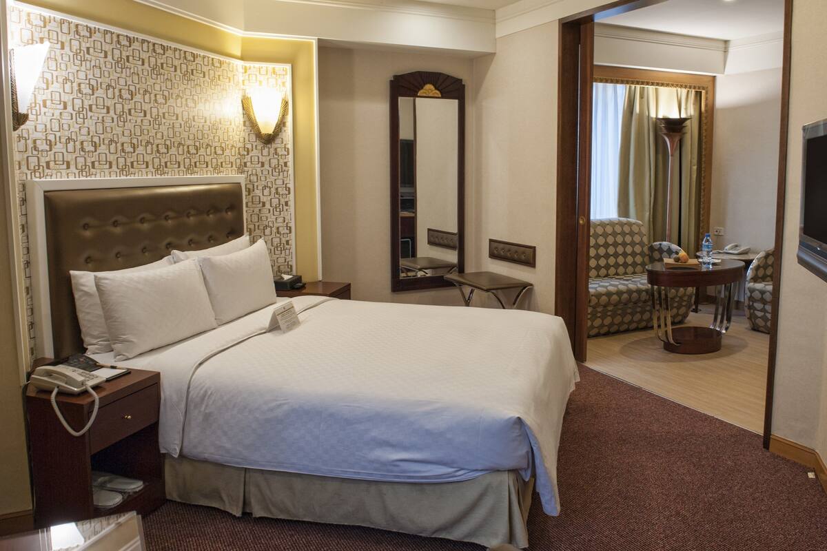 A hotel room with a large bed, a bedside table with a phone, a mirror, a seating area with a table and chairs, and a TV on the wall.