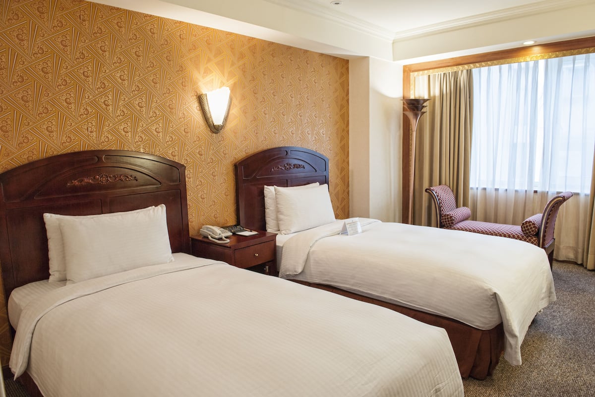 A hotel room with two beds, a nightstand, a lamp, and a window with curtains.